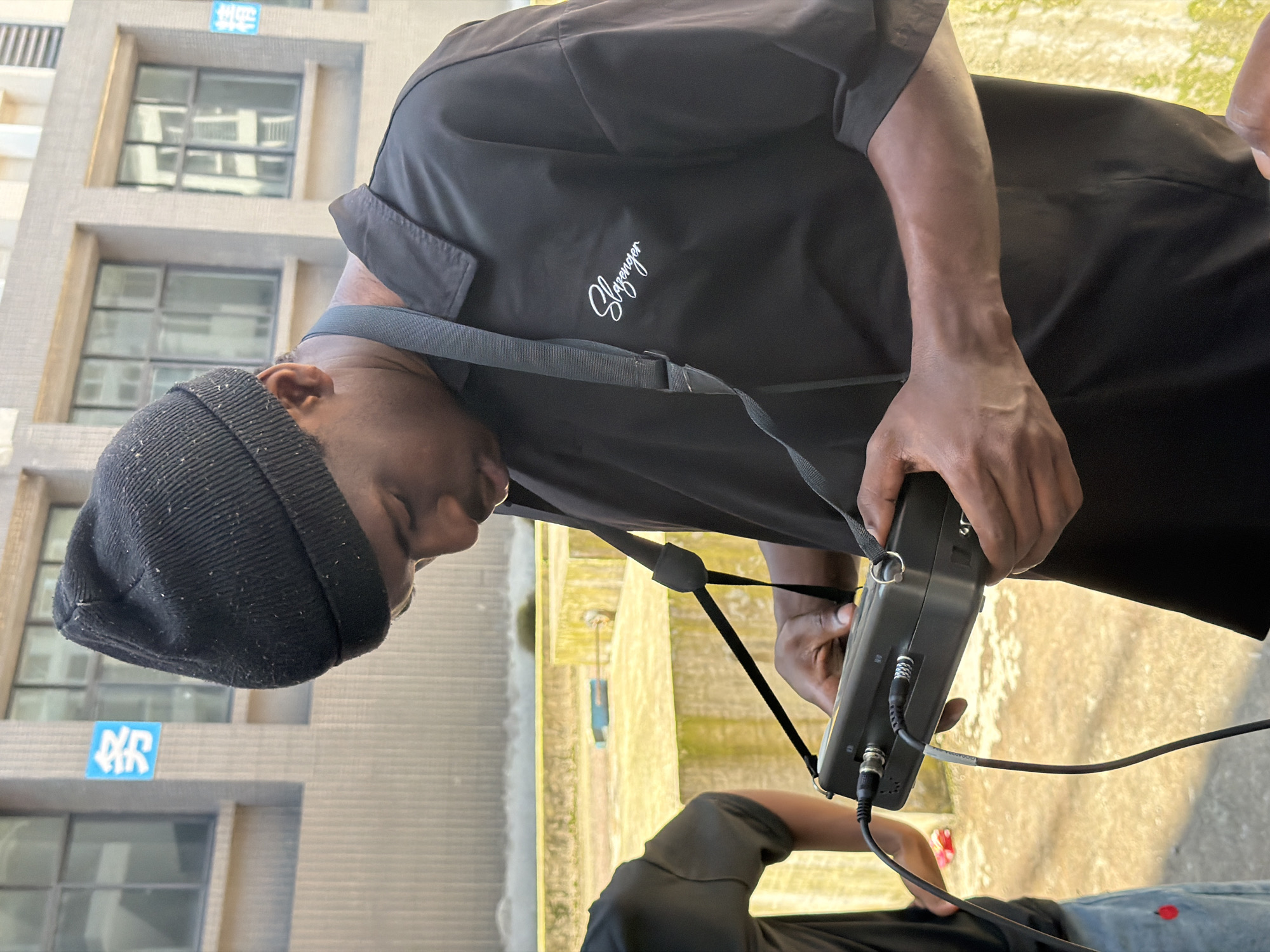 In the city of Jiangjin, China, a young Black man wearing a black beanie and a black collared shirt with the "Slazenger" logo embroidered in white on the left chest, is holding a black electronic device with various cables connected to it. He is looking down at the device, interacting with its controls. To his left, a portion of another person is visible, wearing a black shirt and blue jeans with a red embroidered strawberry. They are standing in an outdoor environment with a beige building in the background, featuring multiple windows. The ground appears to be a concrete surface, possibly a rooftop or patio, with some yellowing and mossy patches. The lighting suggests it is daytime, with sunlight casting shadows.