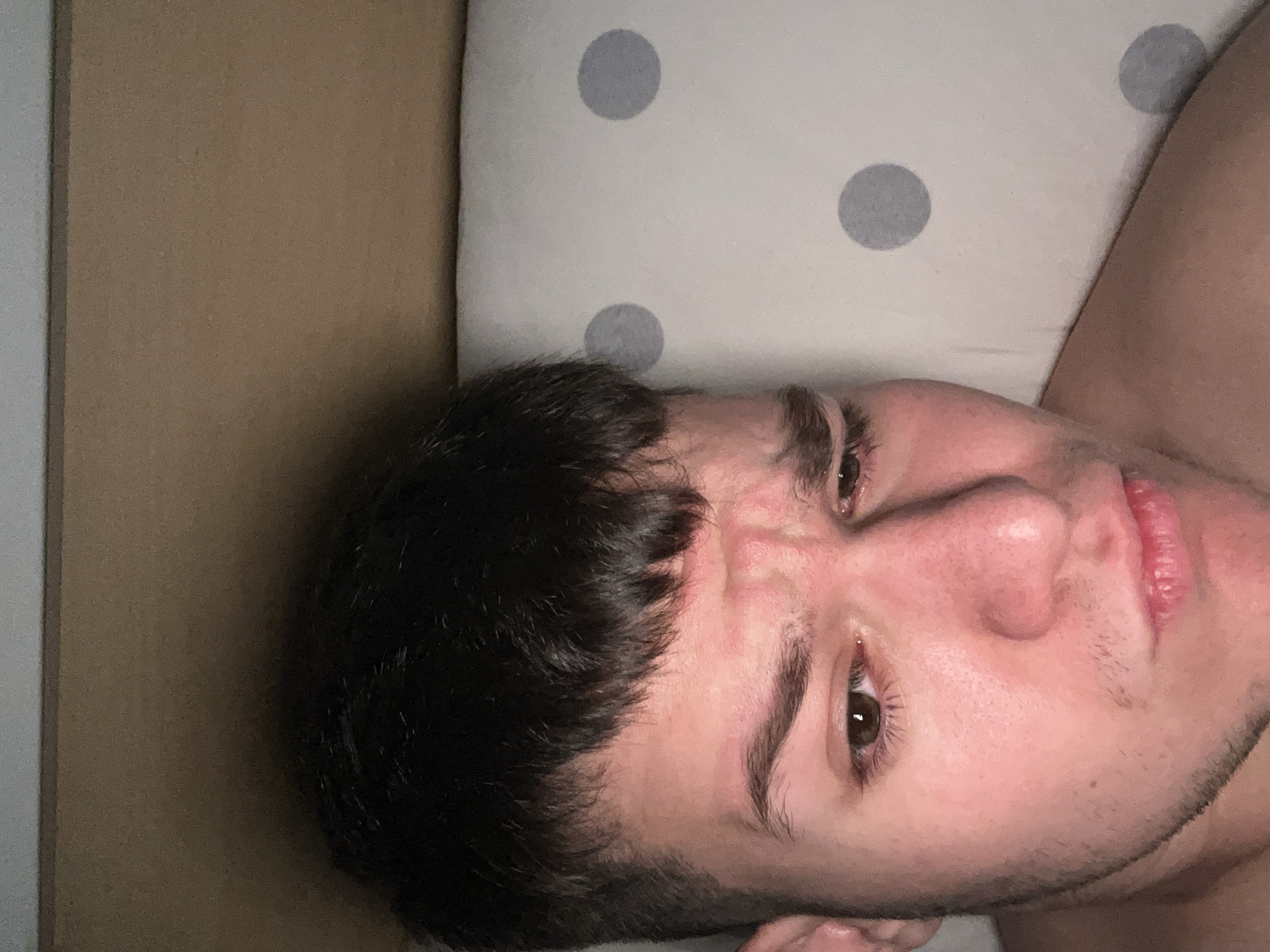 A man is lying on his back with his head on a pillow patterned with gray polka dots. The man has dark hair, brown eyes, and a light complexion. His facial expression suggests discomfort or concentration. He is partially visible from the chest up. To the left of the pillow, there is a cardboard surface. The location is unspecified.
