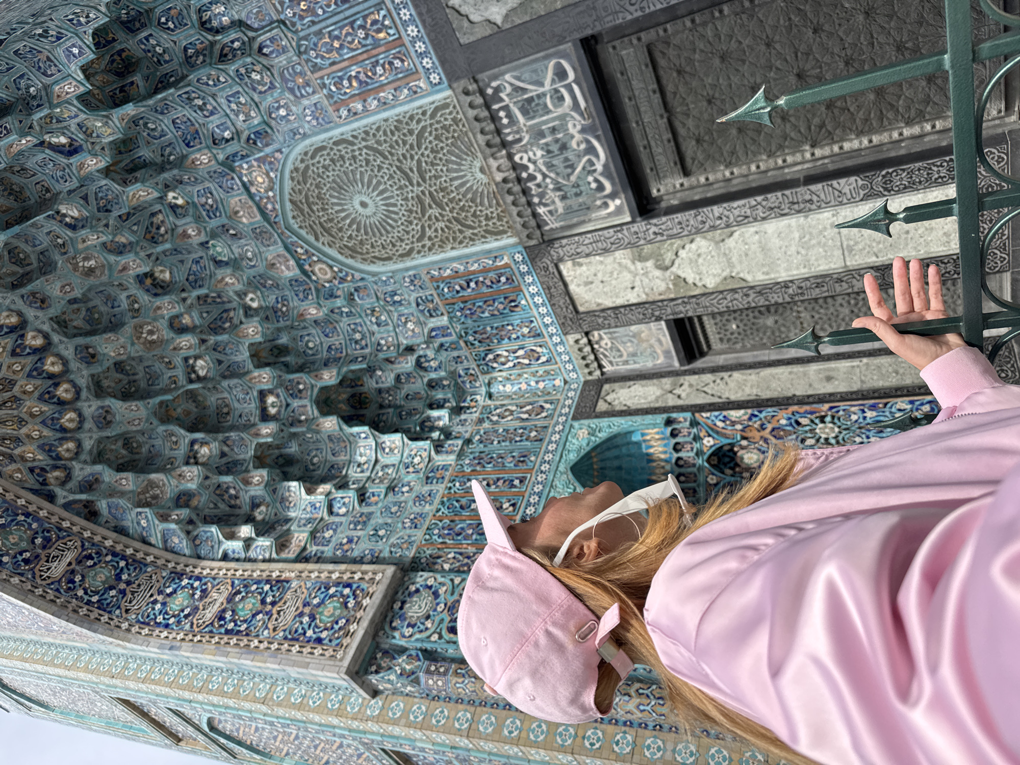 The image captures a vibrant, upward-angled view inside the Saint Petersburg Mosque in Russia, showcasing its magnificent architectural details. The dominant feature is the intricate ceiling and upper wall sections, adorned with dazzling mosaic tiles in various shades of blue, turquoise, white, and gold. These tiles form complex geometric patterns, starburst designs, and subtle floral motifs, reflecting traditional Islamic art. Throughout the tiled surfaces and on darker carved panels, elegant Arabic calligraphy is prominently displayed, likely religious scripture.
In the right foreground, a person is partially visible, looking upwards in apparent admiration of the ornate interior. They have long, light-colored hair and are wearing a pink baseball cap and a lustrous pink satin jacket. A white face mask is pulled down under their chin, and white-framed glasses rest on their head. Their right hand gently holds onto a dark green metal fence or railing, which is embellished with sharp, decorative finials.
The perspective suggests the individual is standing below the grand ceiling, appreciating the intricate craftsmanship. The scene is brightly lit, highlighting the rich colors and detailed textures of the mosaics and carvings. The combination of the human presence and the architectural grandeur conveys a sense of awe and engagement with the sacred and beautiful space.