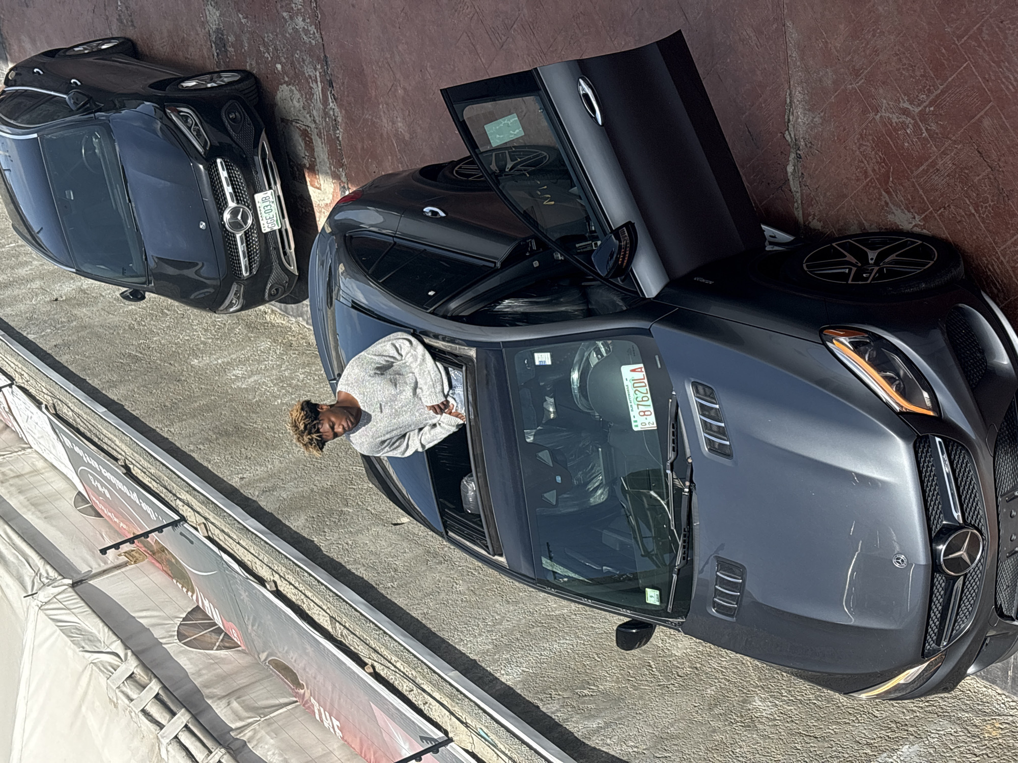 A high-angle shot shows a person in a gray sweater sitting in a dark gray convertible Mercedes-Benz with the door open, parked alongside another dark gray Mercedes-Benz in Igboefon, Nigeria. Both cars have the driver's side facing up and the passenger's side facing down. The convertible has a license plate that reads "08762DLA." A barrier, featuring partially visible advertisements, runs parallel to the vehicles. The ground is concrete, and a red wall is in the background.