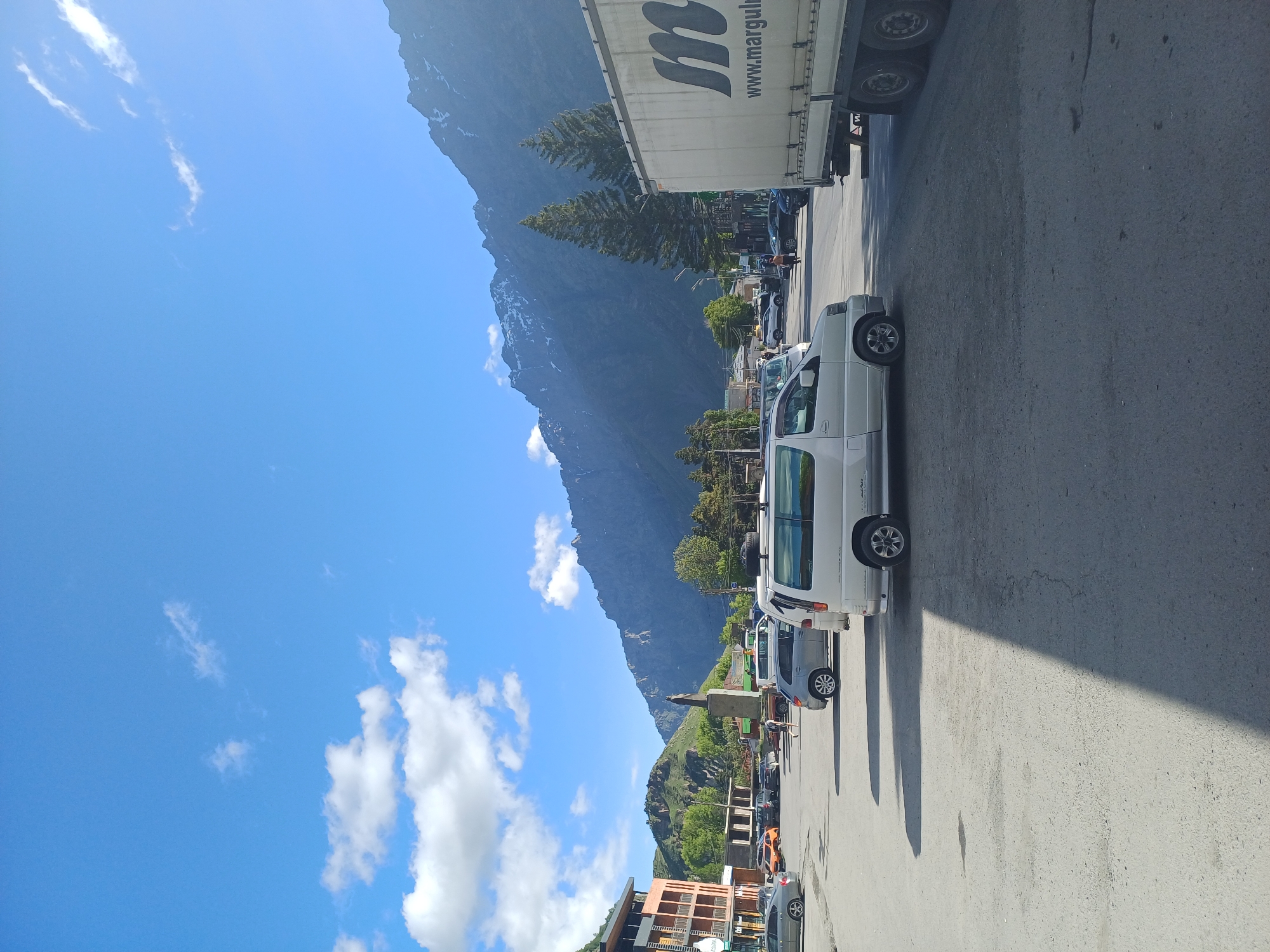 The image depicts a sunlit scene in a town square, likely in Stepantsminda, Georgia, with a prominent backdrop of rugged, snow-capped mountains. The sky is a clear blue with scattered white clouds. In the foreground, a white minivan is parked on a paved surface. To the right, a large white truck with the logo "m www.marguli" is visible, partially obstructing the view. The town square is populated with several other vehicles, including cars and a grey SUV. A building with orange and brown accents stands on the left, and a statue of a person is centrally located in the mid-ground. Green trees dot the landscape, and more buildings are visible in the distance. The overall impression is that of a bustling, somewhat rural, mountain town on a bright day.