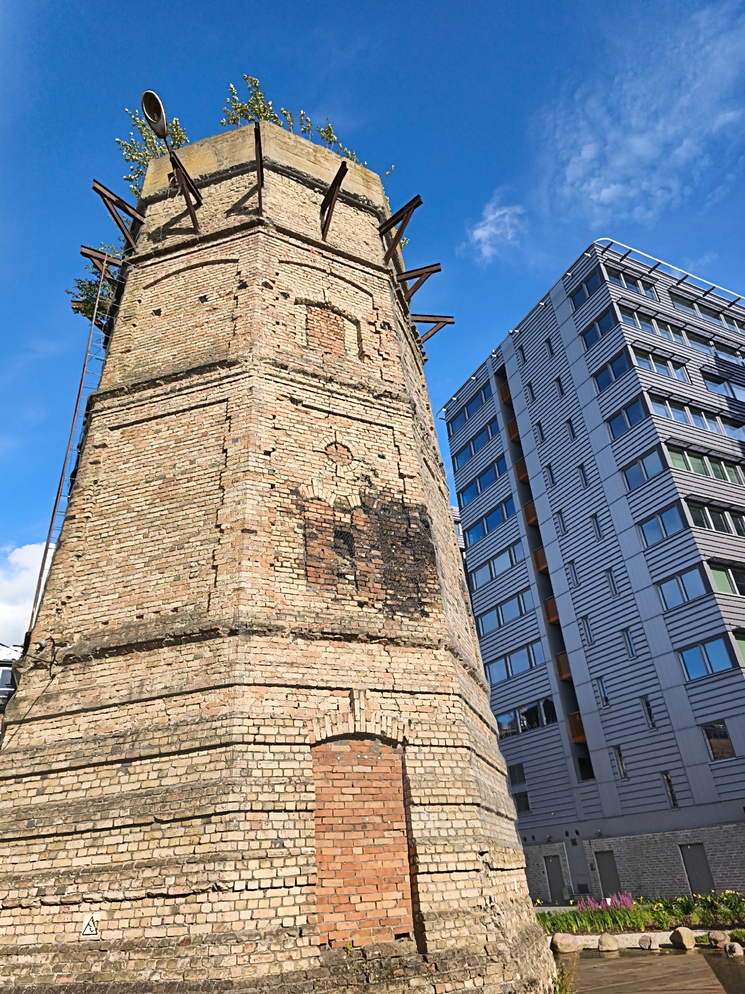 The image shows an old brick industrial structure juxtaposed with a modern apartment building in Tallinn, Estonia. The brick structure is tall and weathered, with visible signs of decay, and is topped with metal supports and some vegetation. A street lamp is mounted on top of it. The modern building, to the right of the brick structure, is a multi-story building with a grey facade and numerous windows. The scene is set against a clear blue sky with scattered white clouds. In the foreground, there is a body of water with decorative rocks and plants, suggesting a landscaped area. The angle of the photograph is low, emphasizing the height of both structures and the contrast between old and new architecture. There are no people visible in the image.