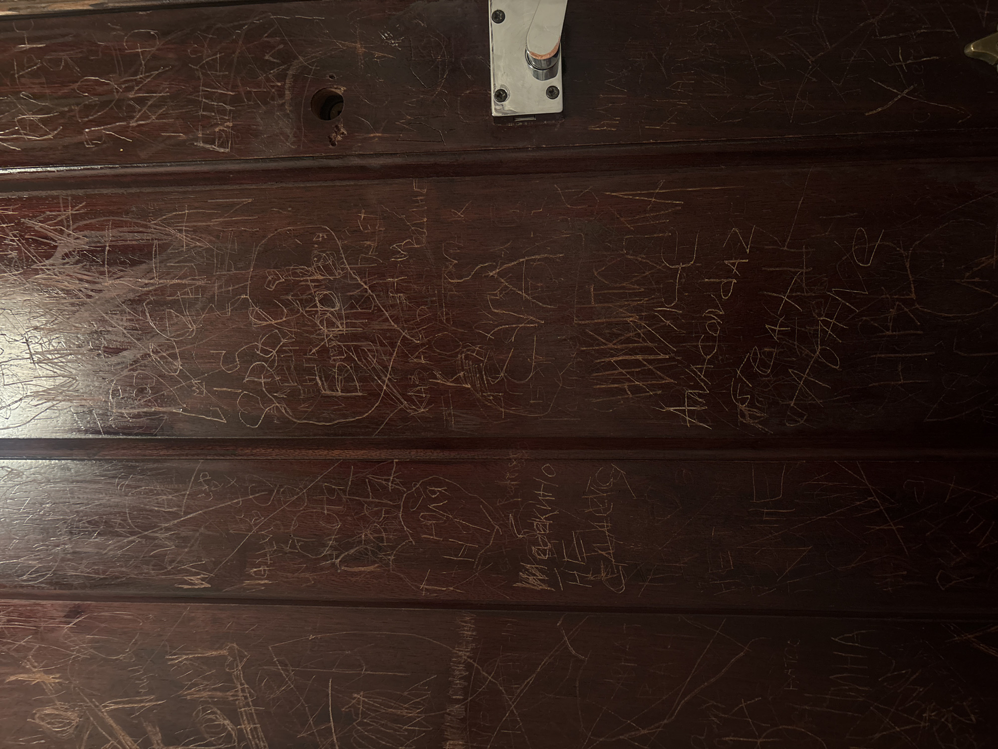 The image depicts a dark brown wooden door panel covered in etched graffiti. The graffiti appears to be mostly names, dates, and simple drawings. A metal latch or handle is mounted on the upper right section of the door. The door is constructed of horizontal panels, indicating it may be part of a train carriage, specifically from a long-distance train from Moscow, Russia.