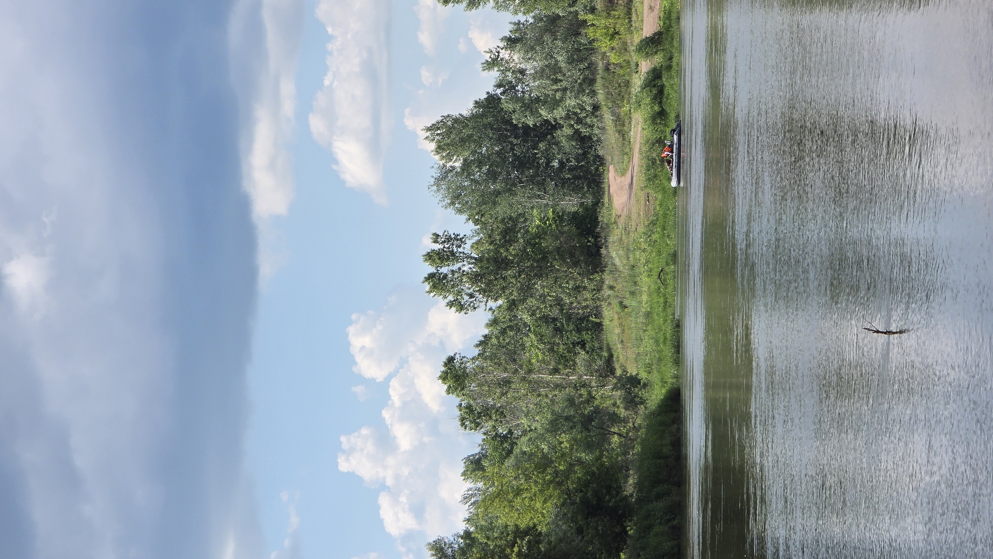 The image shows a wide river with a calm surface reflecting the sky and trees. On the opposite bank, there is a dense line of lush green trees. A dirt path winds through the grass on the bank, leading towards the trees. In the middle of the river, a small inflatable boat with two people in orange life vests is visible. The sky above is partly cloudy, with large grey clouds interspersed with patches of blue sky and smaller white cumulus clouds. The scene appears to be set in a natural, rural environment.