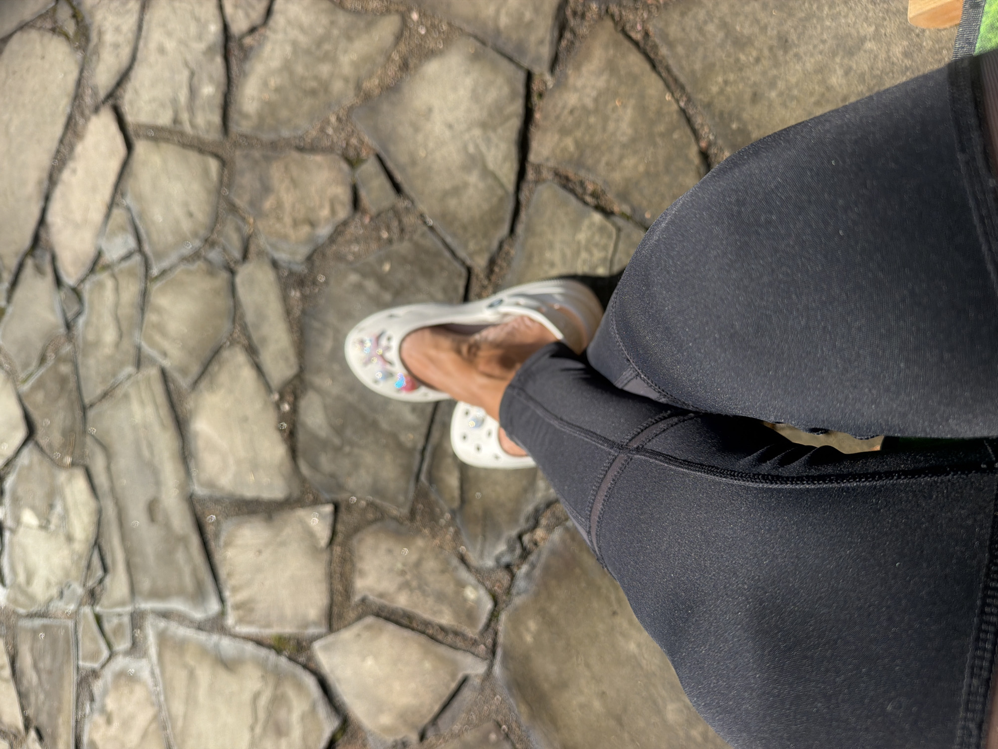 A first-person perspective shot shows a person's legs and feet, wearing dark grey athletic leggings and white Crocs adorned with colorful charms, resting on a sun-drenched patio made of irregular, light brown flagstones. The Crocs are decorated with a floral pattern and small, colorful charms. The leggings feature visible stitching details. The scene is set outdoors, with the stone patio occupying the majority of the frame. There is no indication of the city of Vyazy, Russia.