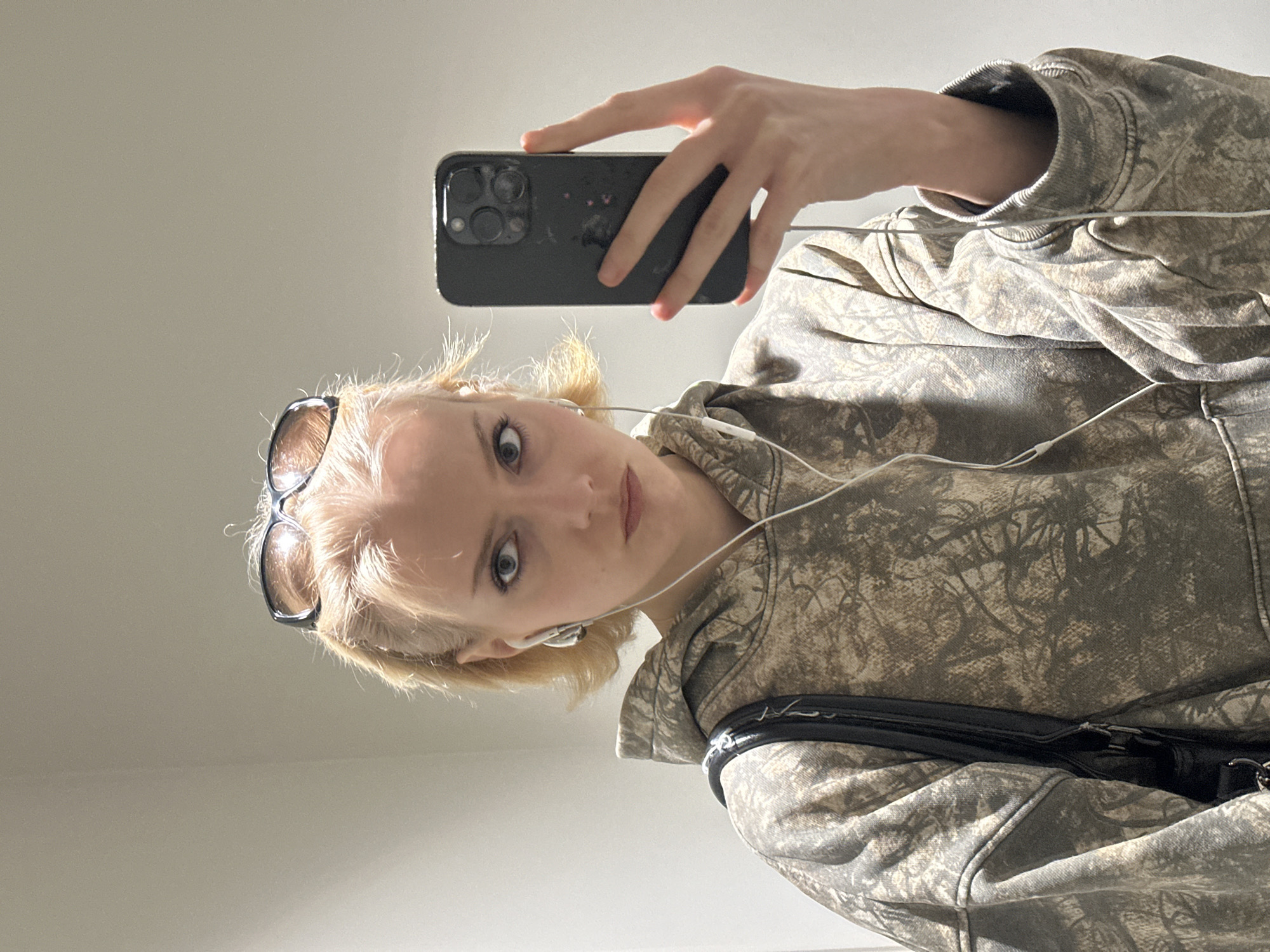 A young woman is taking a selfie with a black smartphone, holding the phone in her right hand. She has short blonde hair and is wearing sunglasses on her head, and earbuds are in her ears. She is wearing a camouflage patterned hoodie with a black shoulder bag. The background is a plain, light-colored wall. There is no information to determine this image was taken in London, United Kingdom.