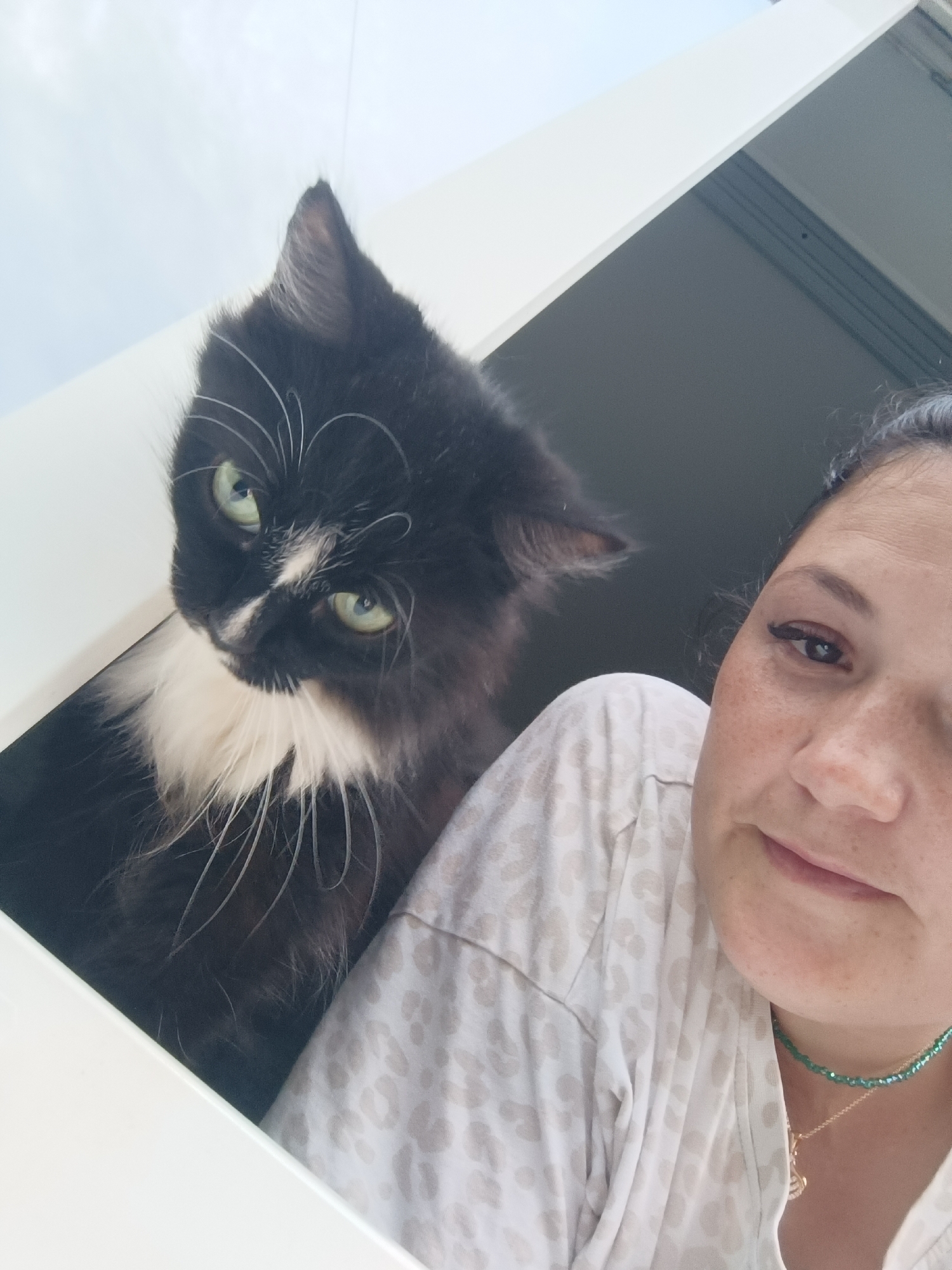 In this close-up, low-angle, outdoor selfie, a woman and a black and white cat are positioned near a window. The woman is on the right, looking towards the camera with her face turned slightly. She has dark hair pulled back, freckles on her face, and is wearing a light-colored shirt with a leopard print pattern and a green beaded necklace. The cat, on the left, is looking at the camera with bright green eyes. It has black fur with a white chest and whiskers. The background consists of a white window frame and a hint of a grey, textured surface. There is no indication of the city of Chaikovsky, Russia within the image itself.