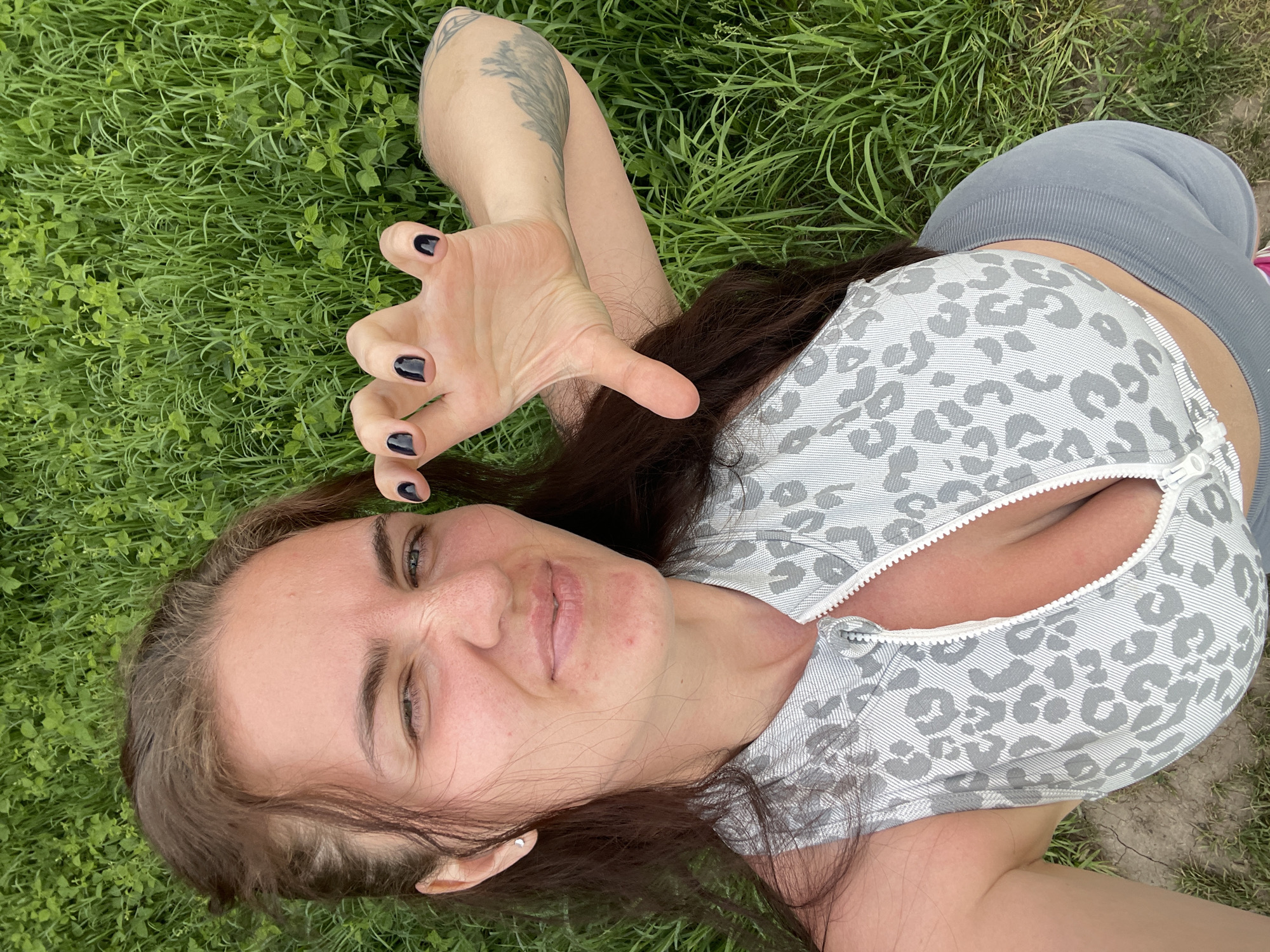 A woman with long, brown hair and dark nail polish lies on a patch of green grass. She is wearing a gray and white patterned top with a zipper and gray leggings. Her left arm is outstretched with her hand positioned to look like a claw. A tattoo is visible on her left forearm. The location appears to be outdoors, possibly a park or grassy area. The city of Ostrov, Russia, is not evident in the image.
