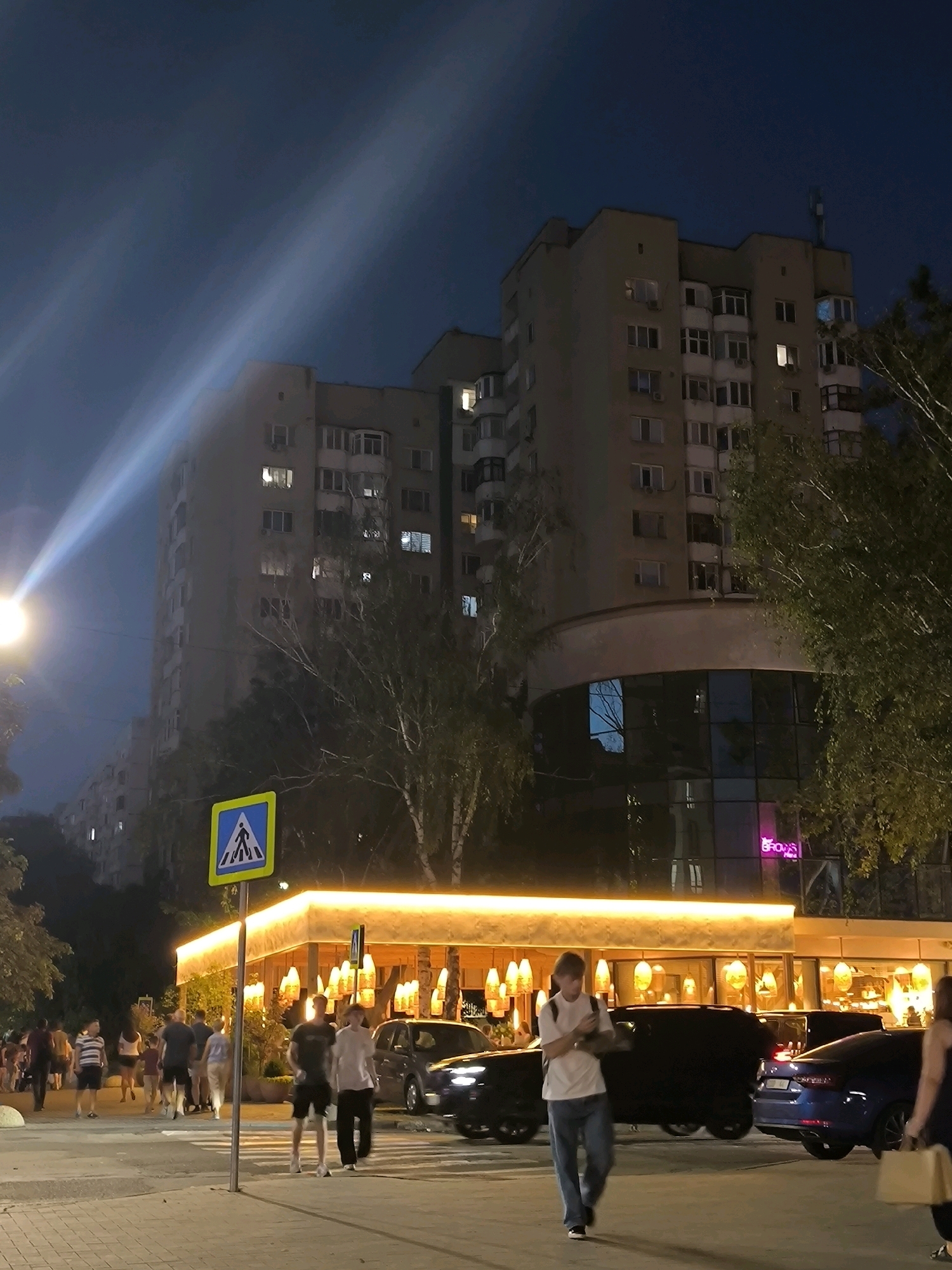 An urban street scene unfolds at night in Chișinău, Moldova, under a dark blue sky illuminated by strong, angled light beams originating from an unseen source, likely a powerful streetlamp or spotlight. In the foreground, a paved pedestrian area leads to a crosswalk marked with white stripes, further bordered by a yellow and blue pedestrian crossing sign on a pole.
Several individuals are present, some walking on the pedestrian path and crossing the street. Two men in the mid-ground are walking across the crosswalk; one wears a dark t-shirt and shorts, the other a light t-shirt and jeans. To the left, a group of people are gathered near the entrance of a brightly lit building.
The background is dominated by a multi-story apartment building with numerous windows, some illuminated. In front of the apartment building, a modern, glass-fronted structure glows with warm interior lighting, featuring hanging decorative lamps. The exterior of this structure is bordered by a luminous orange band. A neon pink sign, partially visible, reads "GROWS". Trees with sparse foliage line the street, adding to the nocturnal ambiance. Several cars are parked or driving through the scene, including a dark SUV and a blue sedan. The overall atmosphere suggests a bustling evening in a residential and commercial area.