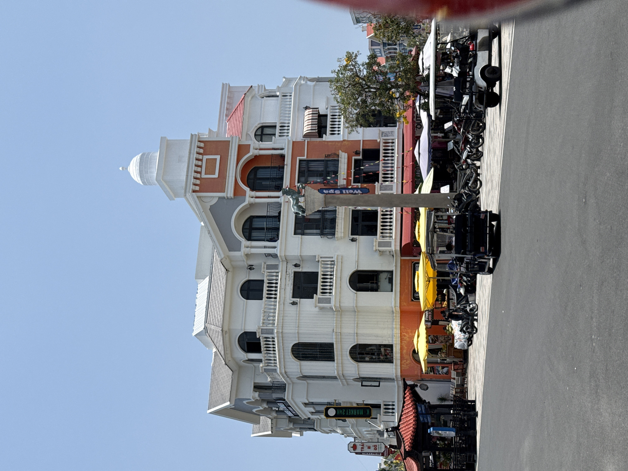 The image captures a vibrant street scene in Phú Quốc, Vietnam, featuring colorful buildings and a sunny sky. The perspective looks up a street, with an array of multi-story buildings dominating the left side, and a paved road stretching into the distance on the right.
The architecture of the buildings is eclectic, with influences suggesting Mediterranean or European styles, characterized by arched windows, intricate white balustrades on balconies, and decorative cornices. Colors are rich, including white, orange/terracotta, and grey facades, topped with red-tiled roofs. One prominent building features a distinctive white dome on its roof, resembling a lighthouse or a grand cupola.
Along the sidewalk, directly in front of the buildings, several motorbikes, scooters, and golf carts are parked, indicating a busy commercial or tourist area. Yellow and white outdoor umbrellas are deployed, suggesting cafes or shops with outdoor seating. A tall street lamp stands in the foreground, adorned with a sculpture that appears to be an angelic figure. The street itself is empty of vehicles and pedestrians.
The weather is bright and clear, with a light blue sky indicating a sunny day, likely morning or early afternoon given the strong shadows.
Several signs with visible text can be identified:
* A blue sign hanging from a pole reads "Well Spa".
* A green rectangular sign on a building states "K-MARKET 24H".
* Below the green sign, another sign reads "EZ STUDIO".
* On a red-roofed building, partially obscured signs include "BIA SAIGON" and "TRÀI BIỂN".
* Other commercial text like "Fresh", "BACARDI", and "OUT" is partially visible on awnings or signs.
No people are explicitly visible in the frame, giving the scene a serene yet inviting atmosphere, hinting at a bustling location anticipating activity.