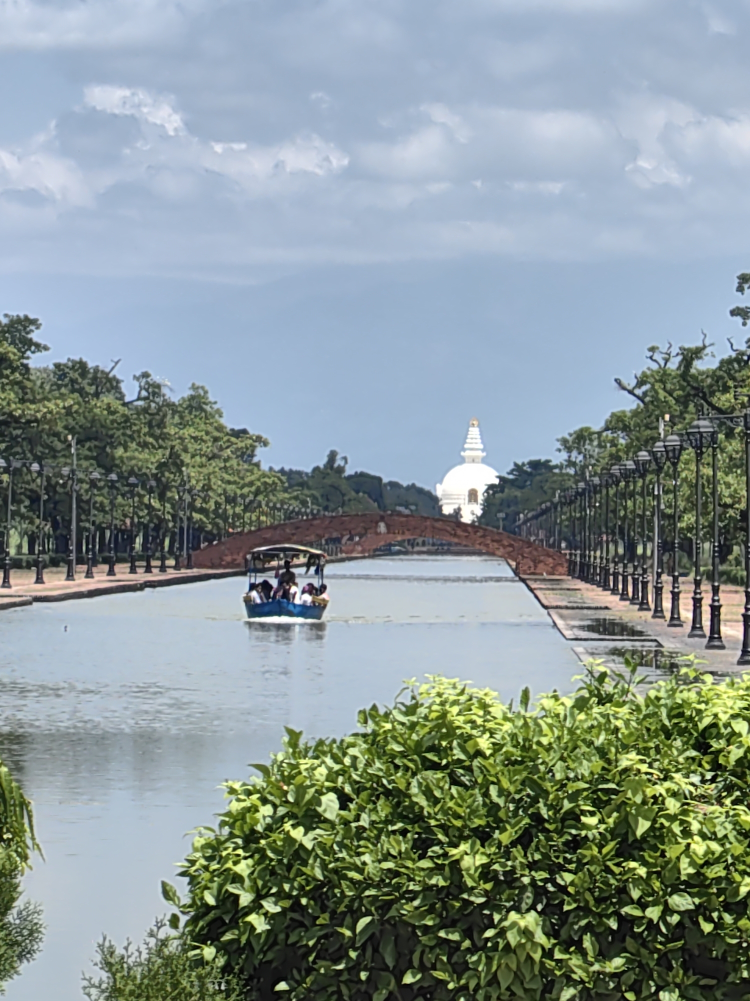 A medium shot captures a scenic view of the Maya Devi Temple pond in Lumbini Bazaar, Nepal, under a cloudy sky. In the foreground, lush green bushes frame the lower portion of the image. The pond dominates the mid-ground, with a blue tourist boat carrying approximately eight individuals navigating its waters. A narrow, arched bridge, constructed of reddish-brown material, spans the pond, leading towards a white stupa with a golden finial, which stands prominently in the background. The banks of the pond are lined with trees and a row of ornamental lampposts. The overall scene depicts a tranquil, tourist-oriented environment.