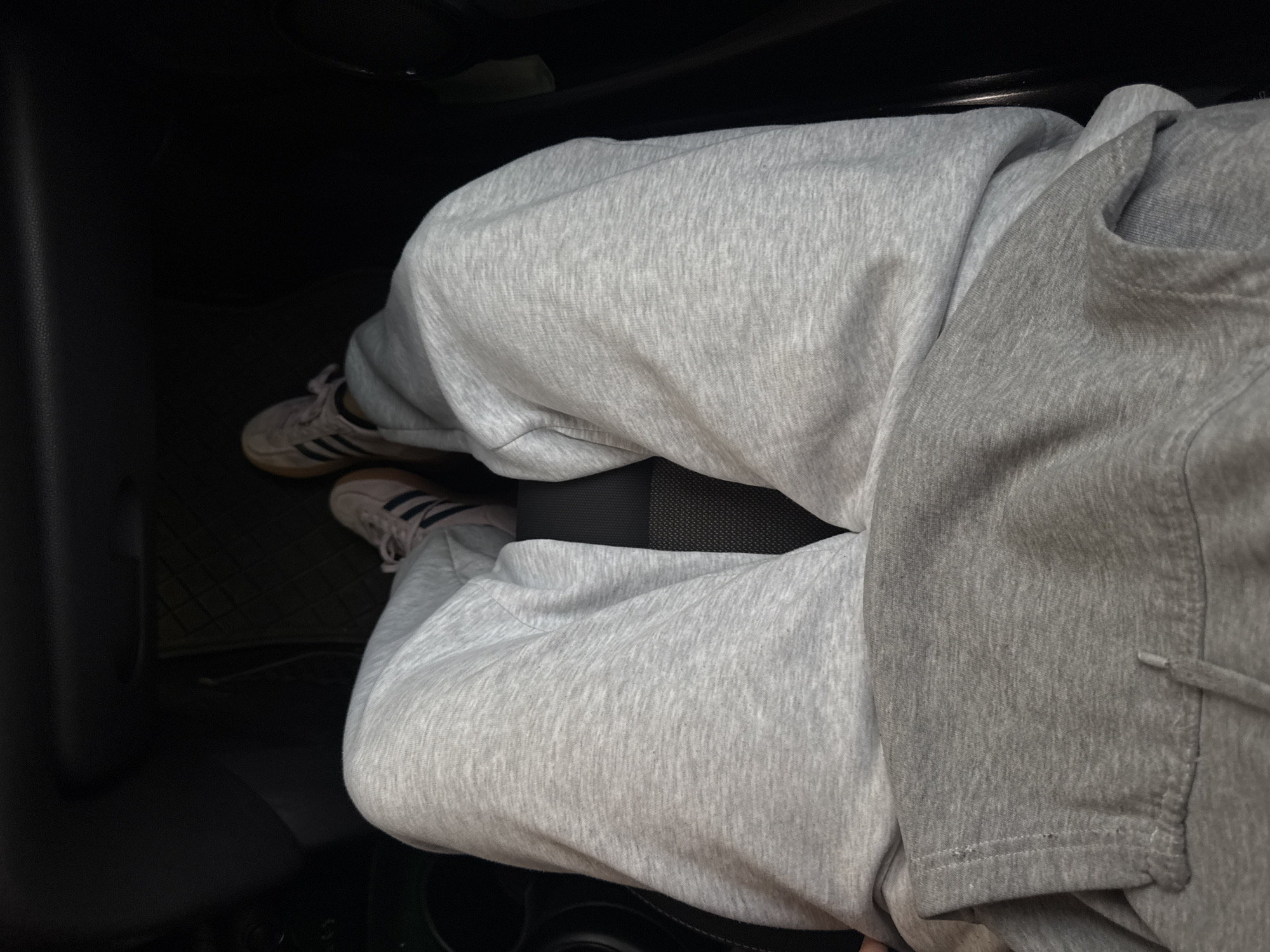 The image shows a person sitting in a car. They are wearing gray sweatpants and off-white Adidas Samba shoes. The car's interior is dark, and part of the car door is visible on the left. The floor mat is green and black. The person is positioned in the driver's seat, seen from an overhead perspective. The photo provides no information about the location in San Francisco Ocotlán (Ocotlán), Mexico.