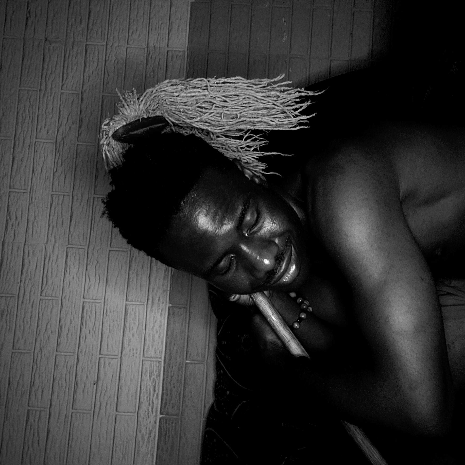 The black and white photograph depicts a dark-skinned individual reclining against a wall with a brick-like texture. The person, who appears to be male, has short dark hair, with a hair covering of thin fiber-like material. They are wearing a beaded necklace and are holding a wooden stick. The person's eyes are closed, and they have a slight smile. The background is mostly dark, obscuring details. The scene is claimed to be in Narayi, Nigeria, but that cannot be verified solely from the image.