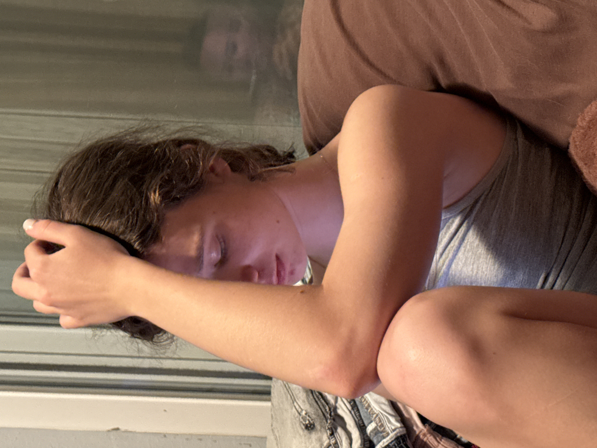 A young person with brown hair is resting their head on their arm. They are wearing a gray tank top and appear to be lying on a bed or couch with a brown cushion. The background is slightly blurred, showing horizontal lines and a possible reflection. The location is unknown.