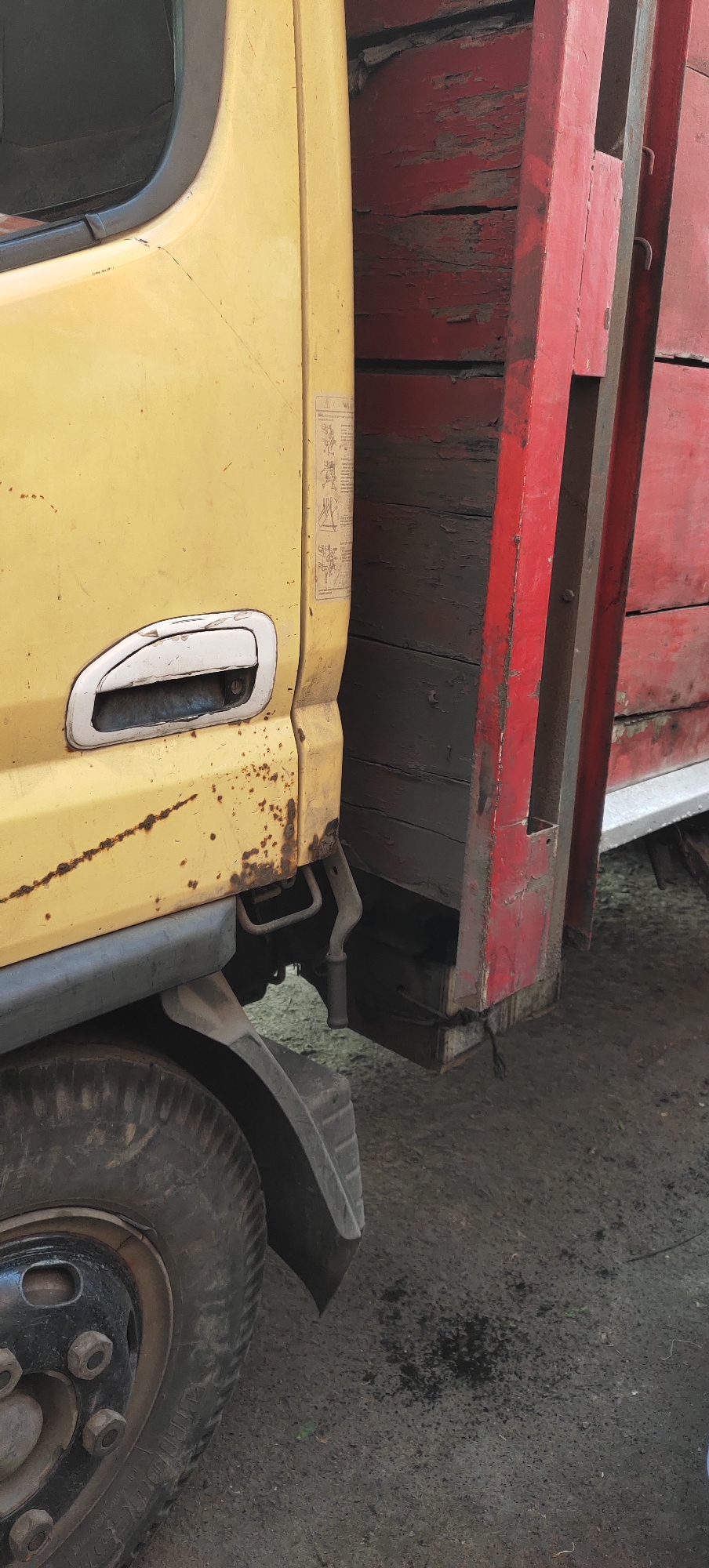 The image shows the side of a yellow truck with a red wooden cargo bed. The truck appears to be parked outdoors on an asphalt or dirt surface. There is some rust on the yellow paint of the truck, and the red paint on the cargo bed is chipped and worn. A black tire is visible on the lower left side of the image. The overall scene suggests a utilitarian vehicle that has seen significant use. No people are visible in the image. The location is identified as Karangtowo, Indonesia.