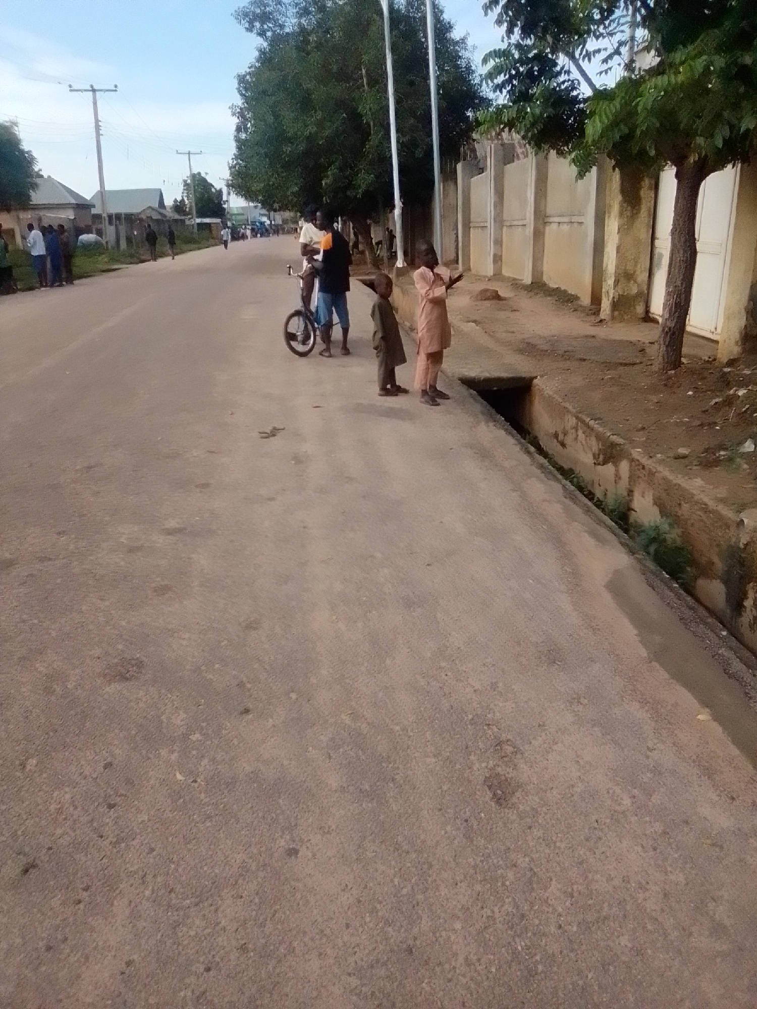 The image depicts a street scene in Gombe, Nigeria, with a paved road and a drainage ditch along the right side. Several individuals are present. In the foreground, two young boys are standing by the road, facing away from the camera. One boy, on the left, is wearing a dark brown outfit, and the other boy, on the right, is in a light brown outfit. Further down the road, a man in a light-colored shirt and dark shorts is standing next to a bicycle, talking to another man in a dark shirt and blue shorts. Other people are visible in the background, some standing on the side of the road near buildings and utility poles. Trees line the right side of the street, and a wall is visible behind them. The sky is partly cloudy, indicating daytime.