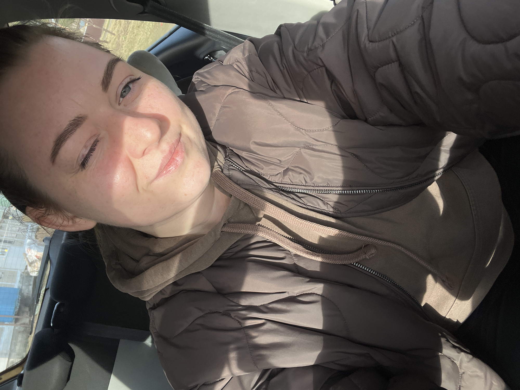 The image shows a woman inside a car. She is wearing a brown hooded sweatshirt and a brown quilted jacket. The location is likely Раздолье, Russia, based on the request. The woman is smiling and winking. Light is shining on her face. The interior of the car is visible in the background. A headrest and parts of the car door and window are visible. Outside the window, some buildings and trees are also visible, although blurry.