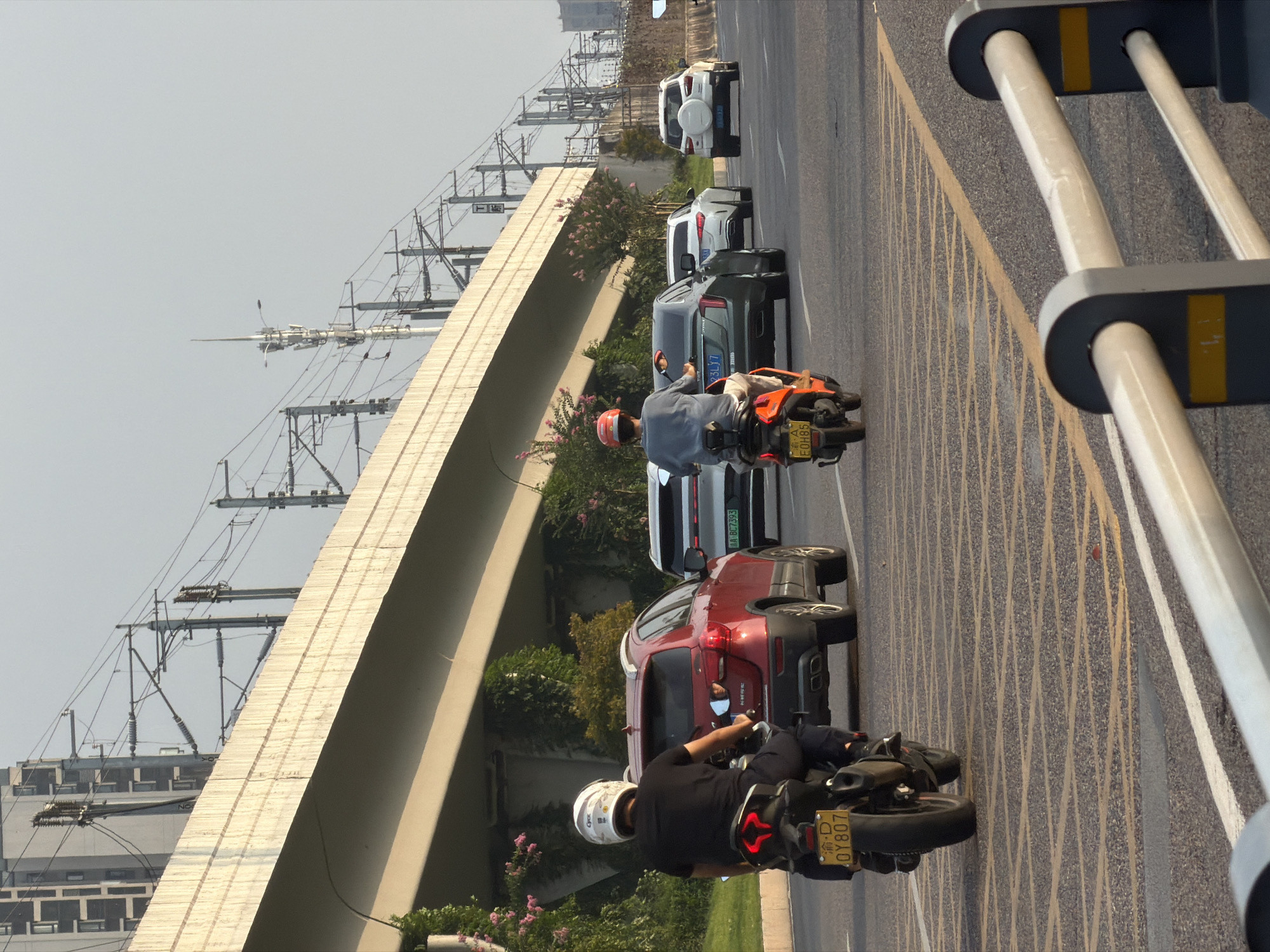 The image depicts a street scene in Jiangjin, China, featuring traffic flow beneath an elevated structure. In the foreground, two motorcycles are visible. The first, on the left, is black with a yellow license plate reading "OY807." The rider, wearing a black shirt and a white helmet, is seated on the motorcycle, facing away from the camera. The second motorcycle, to the right, is orange and black with a yellow license plate that appears to read "EOH85." The rider wears a grey shirt and an orange helmet, also facing away.
Several cars are present in the midground. A red SUV is positioned behind the first motorcycle, its license plate partially obscured. To its right, a dark sedan with a license plate that reads "33LY7" is visible, followed by a white sedan. Further to the right, a white SUV with a spare tire mounted on the back is driving in the same direction.
The environment includes a multi-lane road with yellow and white lane markings. An elevated overpass, constructed of concrete and supported by pillars, dominates the upper portion of the image. Overhead, electrical wires and supporting poles, likely for a railway or power lines, crisscross the sky. Vegetation, including flowering bushes, lines the sides of the road and the base of the overpass. Buildings and other infrastructure are faintly visible in the background against a hazy sky. The lighting suggests it is daytime, possibly late afternoon due to the warm tones.