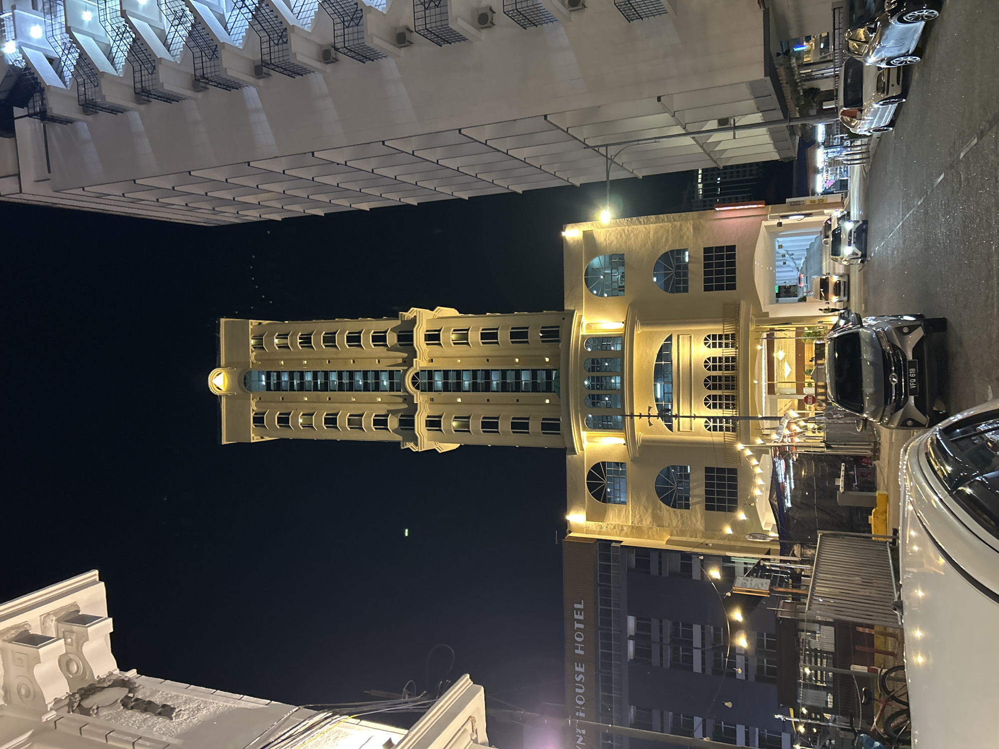 The image shows a street scene at night in George Town, Malaysia, featuring a prominent tall, cream-colored building with many windows and a decorative top, likely a hotel or office building. To the left of this building is a darker, lower building with the text "ONE HOUSE HOTEL" visible on its facade. To the right, a modern white building with external staircases is illuminated. The street is lined with parked cars, and a street light is visible. Several other vehicles are on the road, and there are some string lights and tents set up along the sidewalk, suggesting a commercial or entertainment area. The overall atmosphere is urban and well-lit for nighttime.