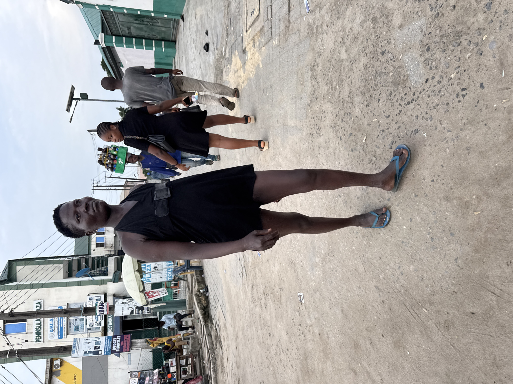 The image depicts a street scene in Lagos, Nigeria, likely in the Ajegunle area, characterized by a mix of commercial buildings and informal market activities. In the foreground, a woman with dark skin and short hair stands facing slightly to the right, wearing a black sleeveless dress and blue flip-flops. Her hands are at her sides, and she appears to be looking off into the distance. Behind her, a woman in a black dress and platform sandals walks away from the camera, carrying a black handbag. Further in the background, a man in a grey t-shirt and light-colored pants also walks away. Other individuals and a vendor with goods on their head are visible in the mid-ground, along with signs for "PENINSULA PLAZA" and "PUBLIC TOILETS." The ground is paved with concrete and dirt, with some debris scattered around. The overall atmosphere suggests a busy urban environment with a pedestrian-focused streetscape.