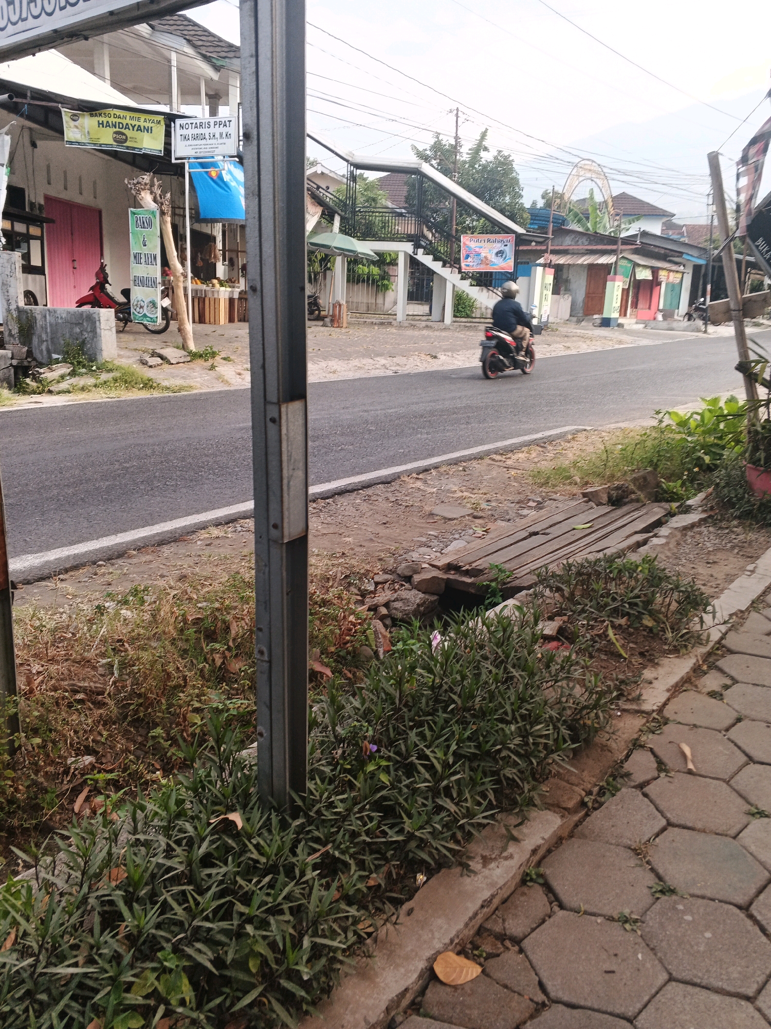 The image depicts a street scene in Ungaran, Indonesia, likely near commercial establishments. In the foreground, a grey metal pole stands vertically, partially obscuring a sidewalk paved with hexagonal concrete blocks. Adjacent to the sidewalk is a patch of dense green vegetation. A narrow, shallow ditch runs parallel to the road, crossed by a makeshift wooden plank bridge.
The road, paved with asphalt, curves gently. On the left side of the road, a building with a white facade and pink doors is visible. A red scooter is parked in front of it. Above the building, a banner reads "BAKSO DAN MIE AYAM HANDAYANI". Another sign indicates "NOTARIS PPAT TIKA FARIDA, S.H., M. Kn.".
Further down the street, on the right side, a person is riding a black and red motorcycle. In the background, several buildings with colorful facades line the street. Electrical wires are strung across the sky. The overall scene suggests a typical urban environment with shops and residential areas. The presence of the "Handayani" banner and "Notaris PPAT" sign indicates specific businesses operating in this location within Ungaran.