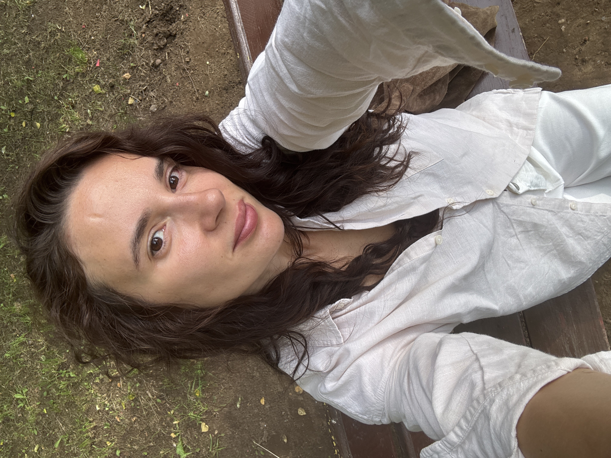 A person is lying down on a wooden bench, facing the camera. Their head is tilted slightly upwards, and they are looking directly at the lens with a soft expression. They have long, dark, wavy hair that is spread around their head. They are wearing a loose-fitting, light-colored button-down shirt, likely linen, and possibly matching pants. The fabric appears textured and slightly wrinkled. The background consists of uneven ground with patches of green grass and dark soil. There are a few small pebbles or debris scattered on the ground. The lighting suggests it is daytime, possibly late morning or afternoon, with natural light illuminating the scene. The overall mood appears relaxed and candid. The setting seems to be outdoors, perhaps in a park or a garden, given the presence of grass and soil. There are no visible signs or text in the image.