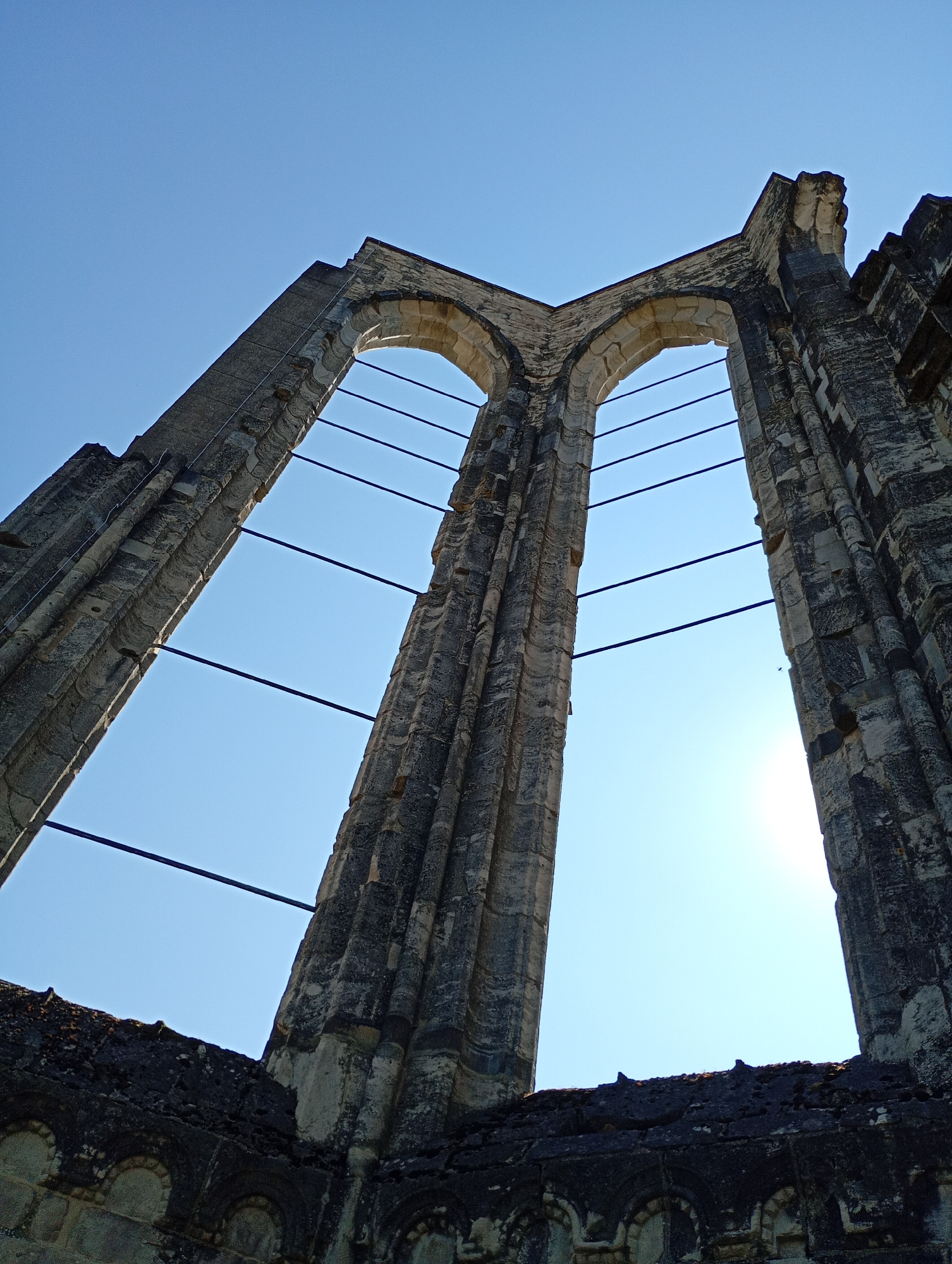 The image shows the ruins of the Walkenried Abbey in Walkenried, Germany, seen from a low angle looking upwards against a clear blue sky. The focus is on the weathered stone arches of a large, gothic-style window opening. The stone is grey and textured, showing signs of age and erosion. Horizontal metal bars are stretched across the opening, likely for structural support or as part of a stabilization effort. The lower part of the image reveals more of the dilapidated stone walls, with remnants of decorative carvings visible on the lower sections. Sunlight shines through the open arches, creating bright areas against the sky. No people are visible in the image.