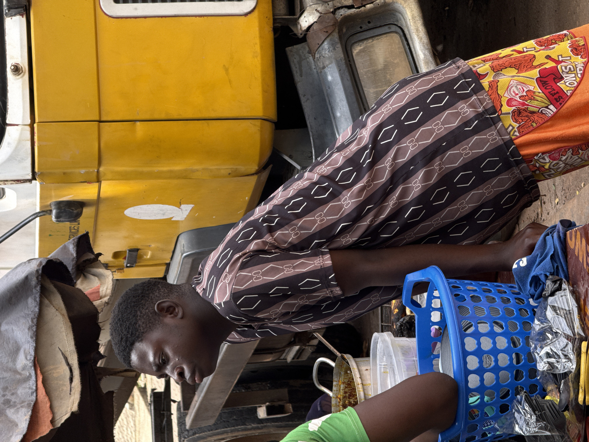 This image depicts a person, likely a young man, in what appears to be an outdoor market or roadside setting in Abuja, Nigeria. He is positioned next to a large, weathered yellow vehicle, possibly a truck. The man is wearing a patterned shirt with a brown and white geometric design and brightly colored patterned trousers, one leg of which is partially visible, showing a yellow fabric with red imagery.
He is leaning over a collection of items, including a blue plastic basket filled with various objects, some of which are indistinct. Next to the basket are clear plastic containers, one of which holds a spoon. The ground appears to be dirt or pavement. The scene is illuminated by natural light, suggesting it is daytime. The overall impression is one of a busy, perhaps somewhat cluttered, environment. The person's expression is not clearly discernible. There is some text visible on the yellow fabric of the trousers, including the word "MONEY" and what appears to be "FLOW". There is also faint white text on the yellow vehicle, too blurry to read.