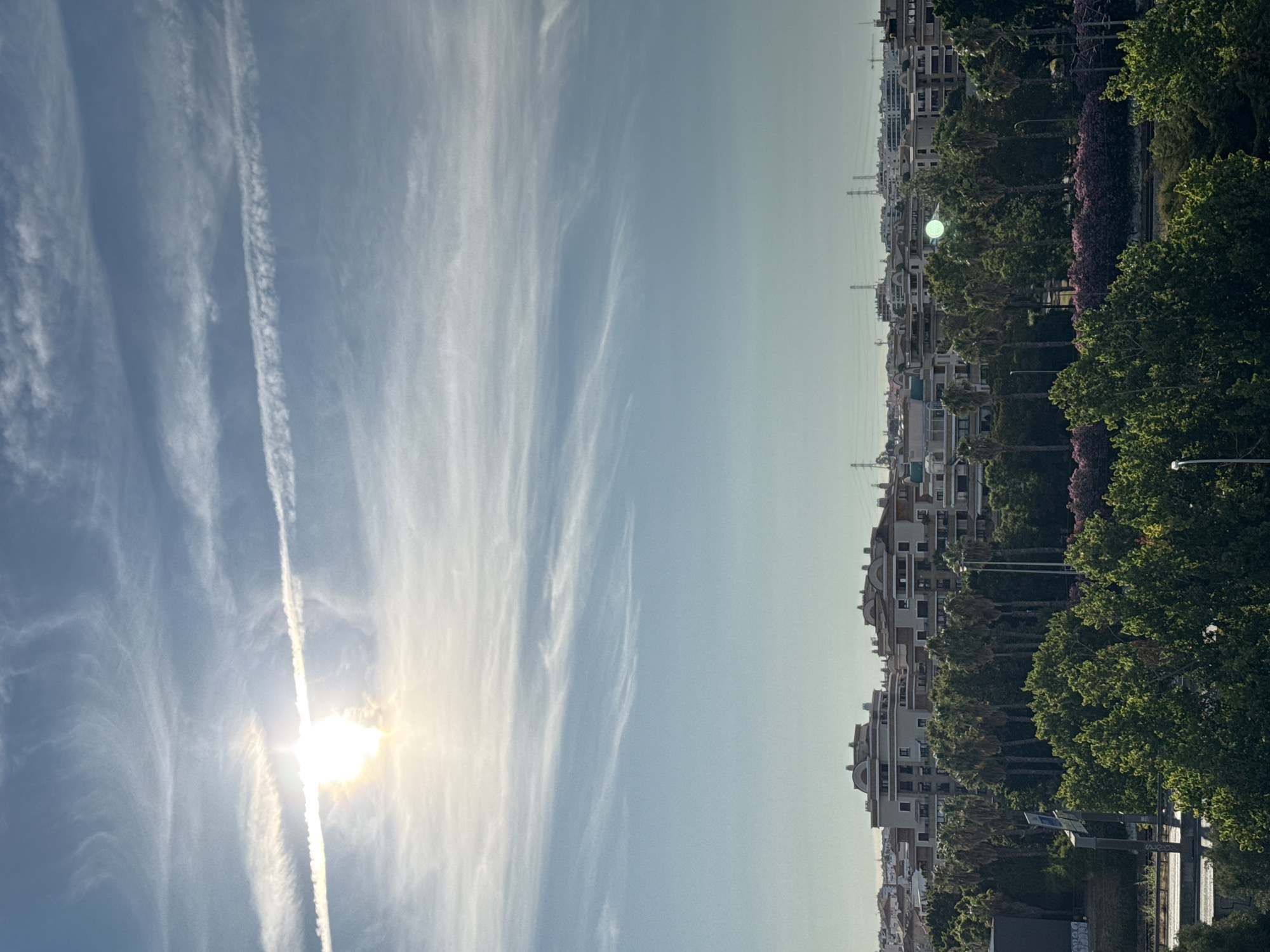 The image depicts a panoramic view of the city of Loures, Portugal, on a bright, partly cloudy day. The sky is a pale blue, with wispy white clouds and contrails from an aircraft stretching across it. The sun, a brilliant white sphere, is partially obscured by clouds, casting a bright glare.
In the mid-ground, a dense line of palm trees and other lush green foliage forms a natural barrier, beyond which a collection of multi-story buildings are visible. These buildings are predominantly light-colored, with pitched roofs and numerous windows. In the distance, a cityscape of more buildings and power lines stretches across the horizon, indicating a developed urban area.
The foreground is dominated by a vibrant green canopy of trees, with hints of pink flowering bushes visible among them. To the left, a concrete barrier or road is partially visible, along with a metal pole and a sign. The overall impression is of a sunny day in an urban environment with significant greenery. There are no people or animals clearly visible in the frame.
