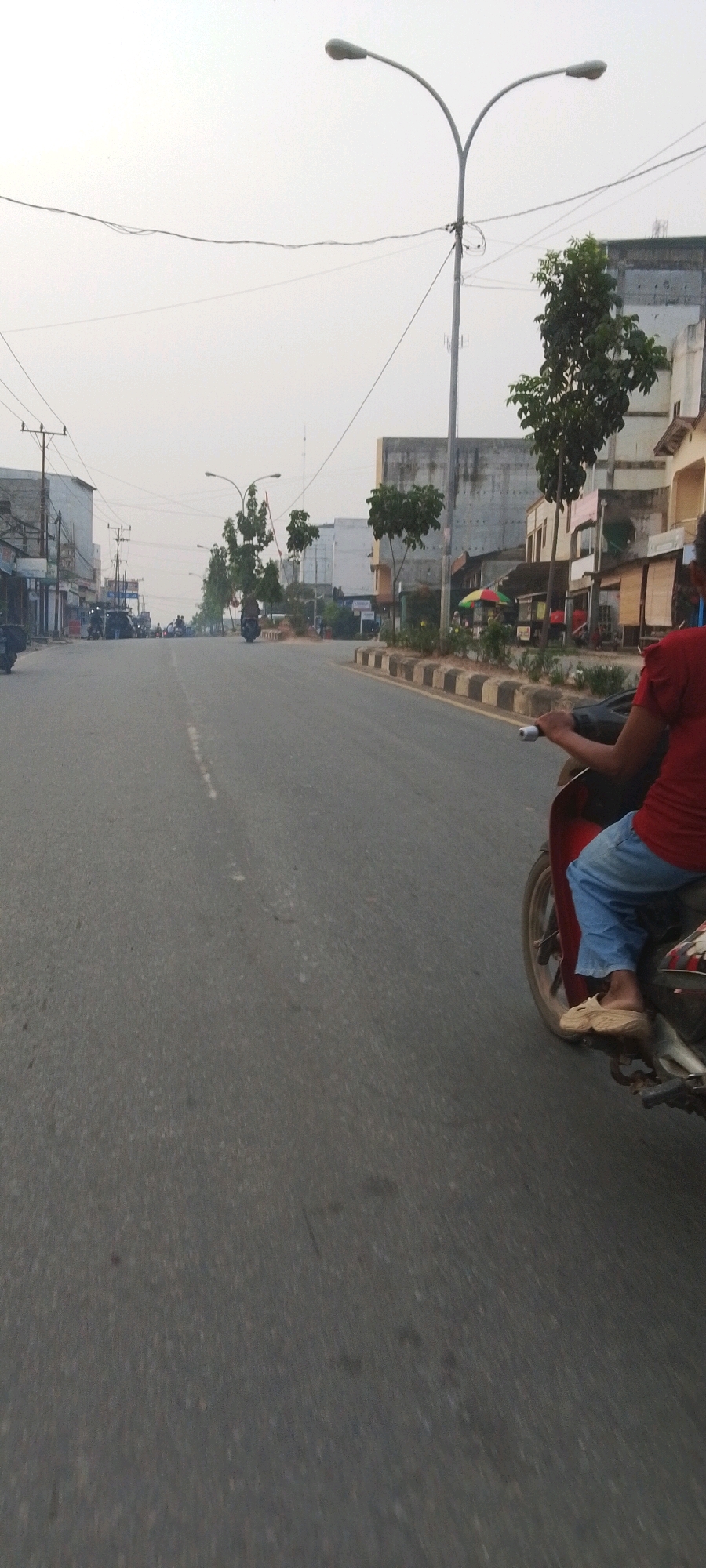 The image depicts a street scene in Lenggadai Hulu, Indonesia, captured from the perspective of someone riding a motorcycle. The road is asphalt with a faded white line marking the center. On the right side of the road, a person is riding a red motorcycle, wearing a red t-shirt and blue pants. They are wearing light brown sandals. The motorcycle has a dark colored rear fender and a sticker on the side. The person's hands are on the handlebars. The road is lined with buildings on both sides. On the right, there are shops with colorful awnings and some trees. On the left, there are more buildings, some with exposed electrical wiring and poles. Streetlights are visible along the road. The sky is overcast with a hazy appearance. In the background, the road recedes into the distance, with other vehicles and people visible, suggesting a moderately busy street. The overall scene is typical of a street in a town or city in Indonesia.