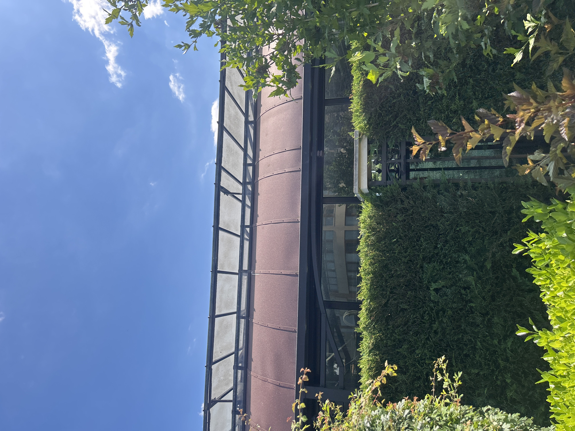 The image, taken in Varna, Bulgaria, captures an exterior view of a building with a curved, metallic facade, possibly a cylindrical section, made of a reddish-brown material with visible horizontal banding. This section is framed by a dark metal structure that supports panels of frosted or translucent glass on the left. To the right of the metallic facade are large windows reflecting the sky and surrounding greenery.
The building is extensively covered and softened by dense, dark green hedges and lush trees with bright green and reddish-brown leaves, suggesting a well-maintained urban landscape or garden setting. Some of the foliage hangs into the frame from above and the sides, creating a natural, almost overgrown appearance. A black metal railing or fence is partially visible between the hedges.
The sky is a clear, vibrant blue with scattered white clouds, indicating a sunny day. The lighting suggests it is daytime, likely late morning or afternoon, with the sun casting shadows and highlighting the textures of the leaves and building materials. There is no visible text in the image. The scene evokes a sense of tranquility and nature integrated with modern architecture.