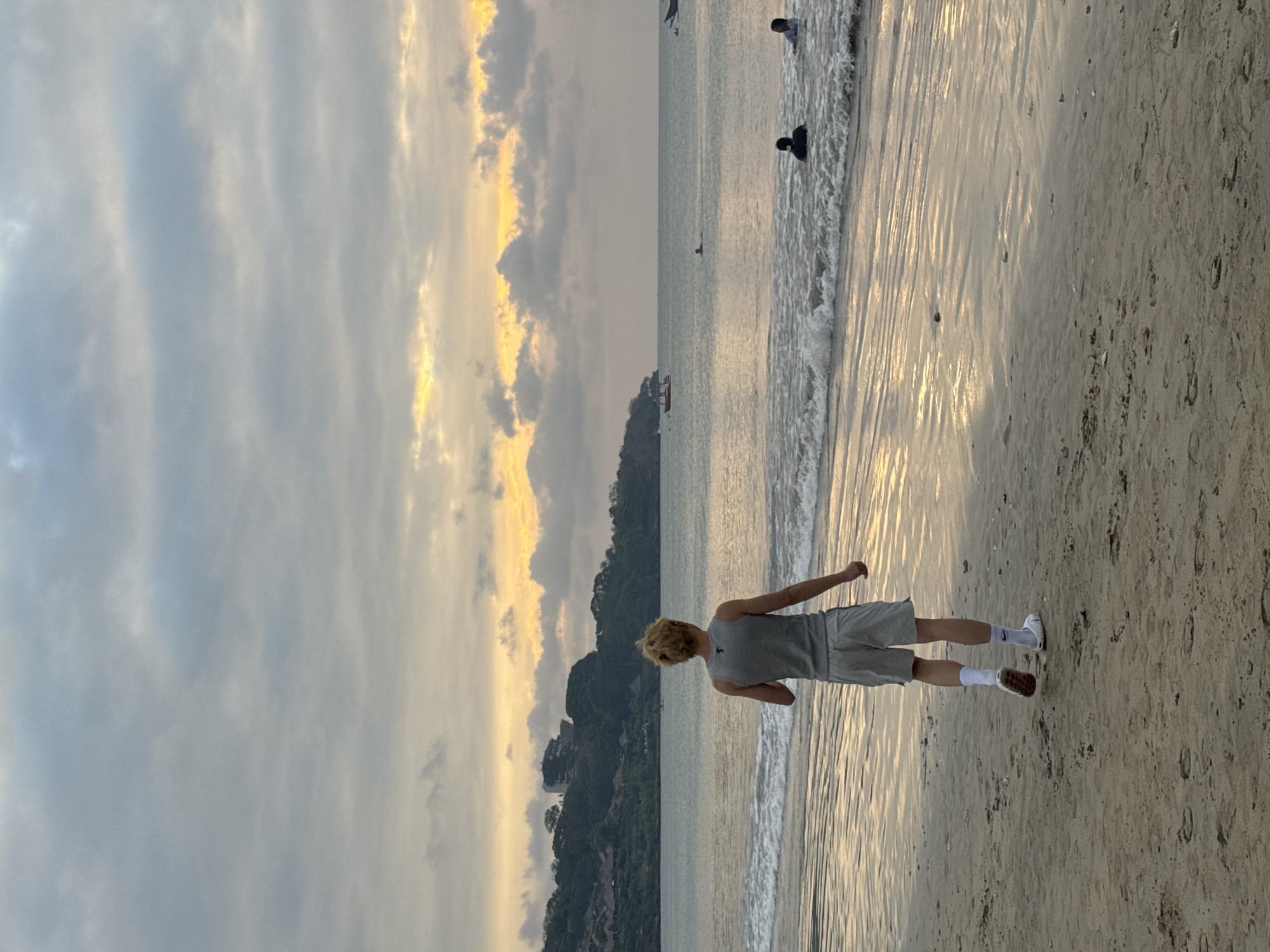 A young blonde individual stands on a sandy beach facing the ocean. They are wearing a grey tank top and light grey shorts, white socks, and brown shoes. The person's left arm is extended slightly. The scene depicts a beach at what appears to be sunset, with the sun casting a warm glow on the water and clouds. The sky is a mix of grey clouds with streaks of yellow and orange from the setting sun. The ocean has gentle waves rolling onto the shore. In the distance, a lush green island with some buildings is visible. Other people are in the water and on the shore further down the beach. The overall mood seems peaceful and serene.