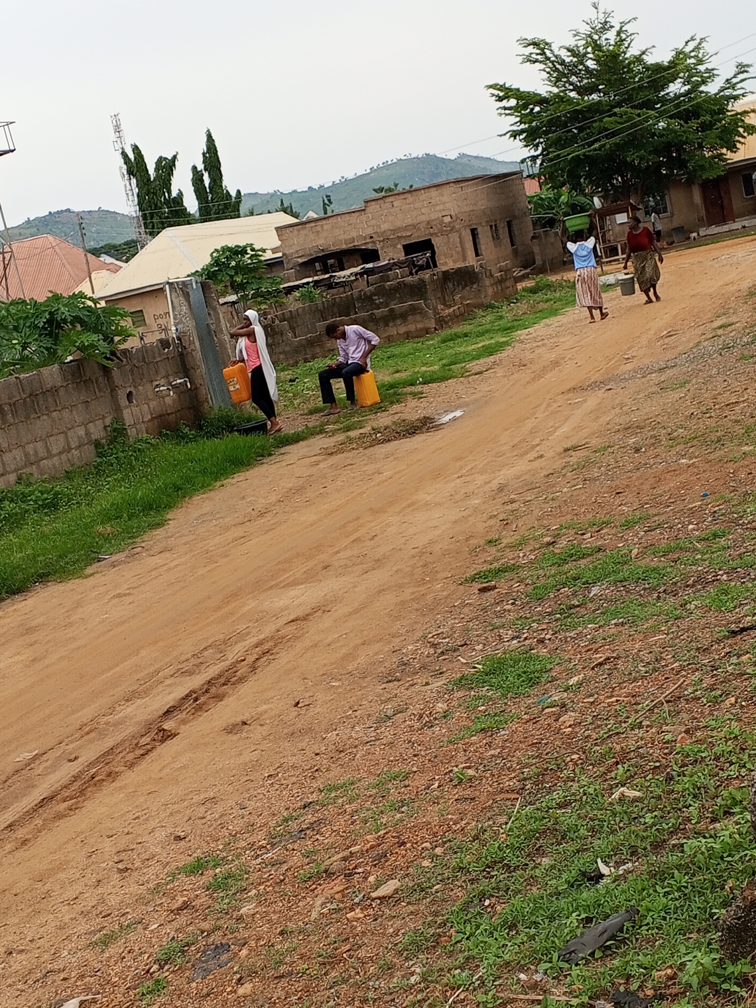 In Ashata, Nigeria, an outdoor scene depicts several individuals engaged in various activities. A person wearing a white hijab and a pink top stands near a water source, holding an orange jerrycan. To their right, a young man in a light purple shirt and dark pants sits on a yellow jerrycan. Further in the background, two women are walking, one carrying a green bowl on her head and the other holding a bucket. They are on a dirt road that winds through a residential area with brick buildings and a large tree. The environment appears dry, with scattered green vegetation and a cloudy sky.