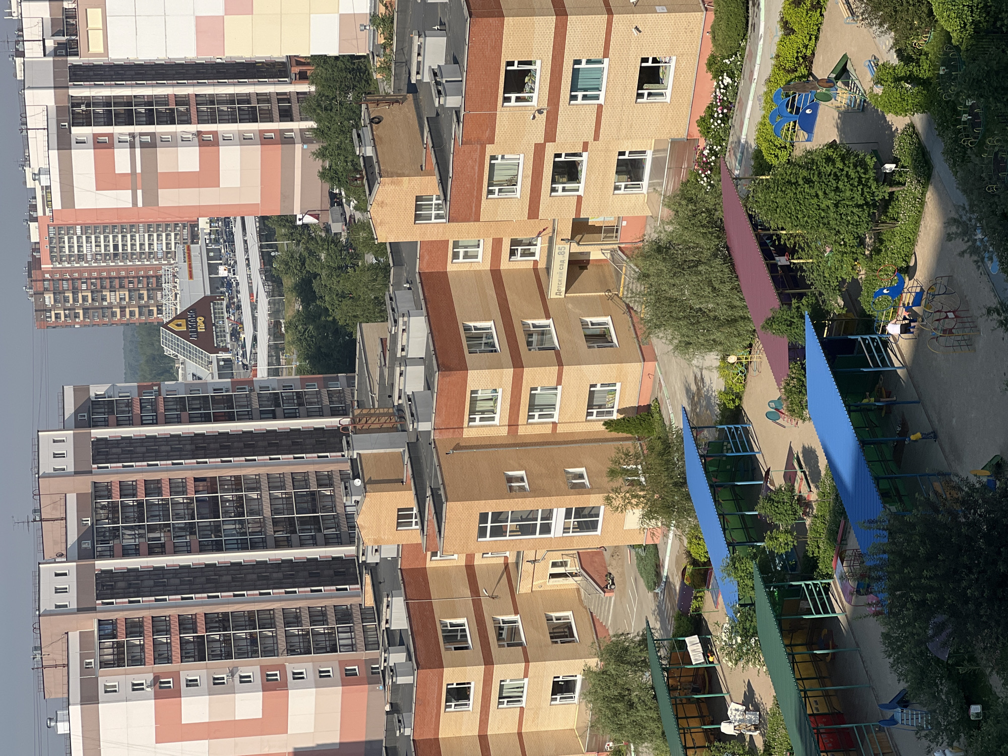 The image shows a residential complex in Novosibirsk, Russia, with several multi-story apartment buildings and a kindergarten or playground area. In the background, tall, modern apartment buildings with colorful facades dominate the skyline. In the middle ground, a brick building with orange and red accents, identified by a sign as "Детский сад 85" (Kindergarten 85), is visible. In the foreground, a playground area with covered sandboxes, slides, and swings is set up. There are a few individuals present on the playground, some children and adults, engaged in recreational activities. The environment appears to be a sunny day with clear skies.