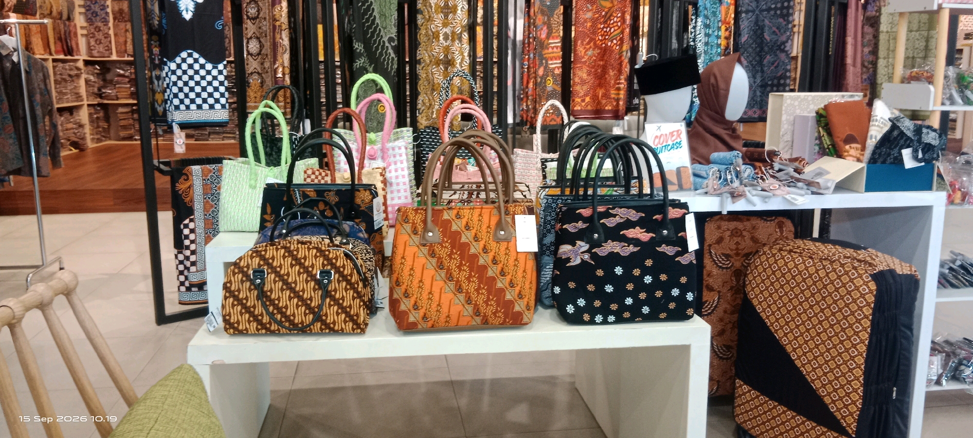 The image depicts a retail display of batik bags and fabrics inside a store. The items are arranged on white shelving units and a rack. In the foreground, several batik handbags are showcased, featuring various patterns and colors such as orange with geometric motifs, black with cloud and bird designs, and brown with intricate traditional patterns. Behind these, a selection of woven bags in pastel colors like green and pink are visible. The backdrop consists of shelves filled with rolled batik fabrics and garments displaying colorful patterns. A mannequin head wearing a black peci (a traditional cap) and a brown hijab is positioned on the right side of the display, next to a sign that reads "COVER SUITCASE". The store appears to be well-lit and organized, with wooden flooring and shelves visible in the background. The overall scene suggests a commercial environment specializing in Indonesian batik products, likely located in Surakarta, Indonesia, a city renowned for its batik heritage.
