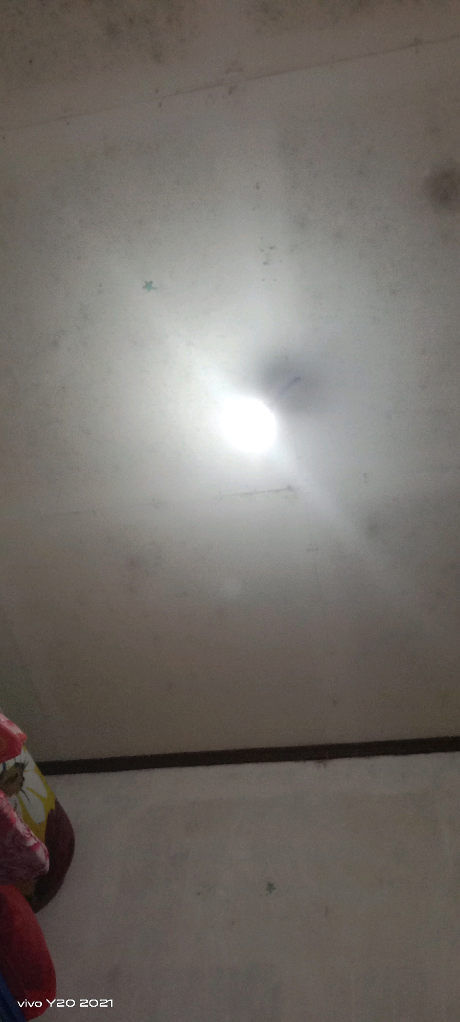 The image shows a ceiling with some dark spots and a bright light source in the center. There is a thin dark line running horizontally across the lower part of the image, which appears to be the edge of a wall or a shelf. To the left, a portion of what looks like fabric with a floral pattern is visible. The timestamp "vivo Y20 2021" is present in the bottom left corner. The scene appears to be indoors, likely a room in a residential setting, and no specific location in the city of Unknown, Indonesia can be determined from the visual information provided.