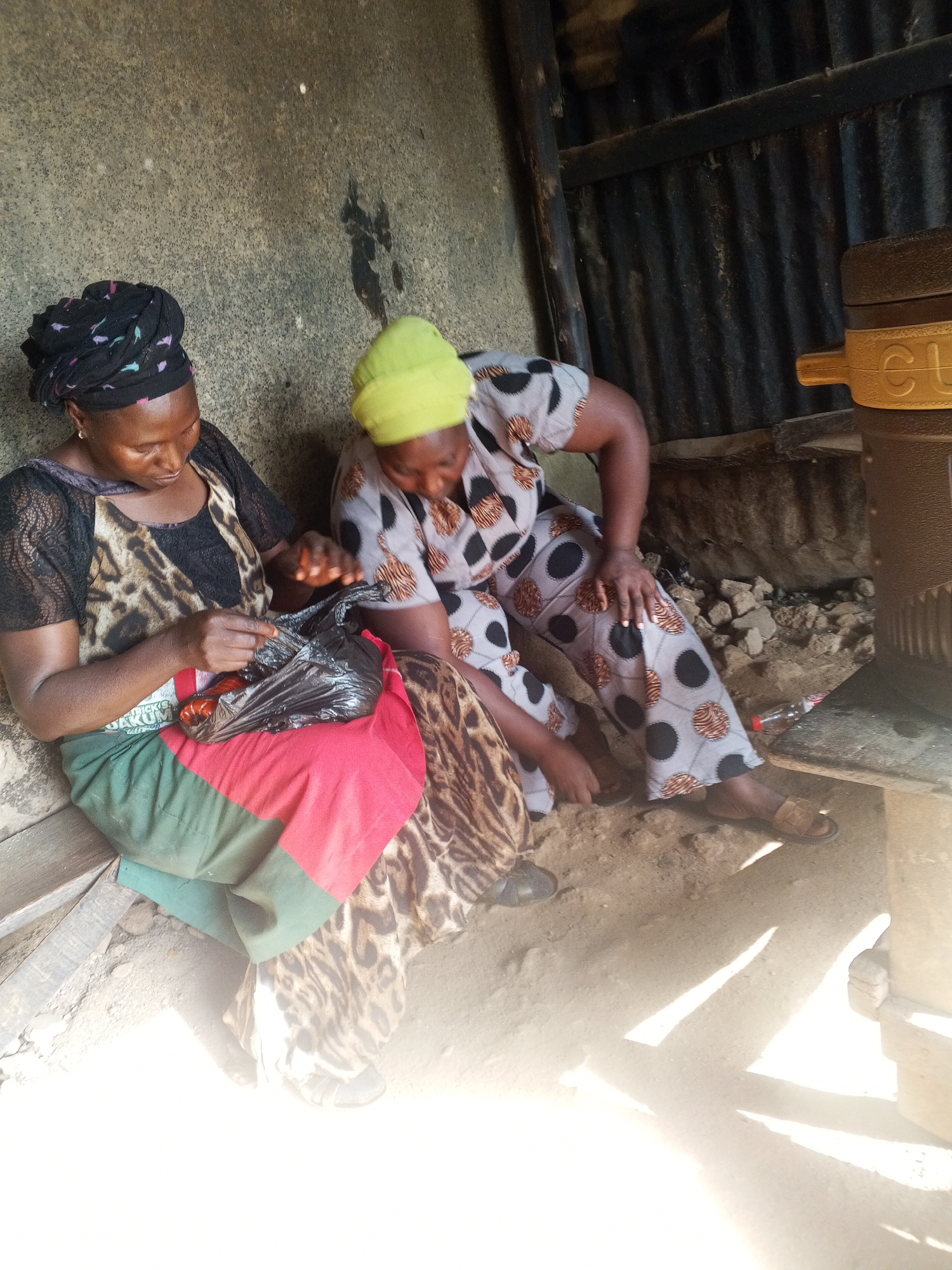 Two women are seated on the ground in a rustic, outdoor setting. The woman on the left, wearing a black headscarf and a patterned dress with a leopard-print apron, is holding a black plastic bag containing what appears to be food items. She is focused on the contents of the bag. The woman on the right, wearing a yellow headscarf and a patterned dress, is also looking down, her hands resting on her lap. They are sitting next to a rough, textured wall and a corrugated metal structure. The ground is sandy and uneven, with some scattered debris. Sunlight streams in from the right, casting long shadows. A large, cylindrical object with yellow accents is visible on the right side of the image. The overall impression is one of a humble, everyday scene.
