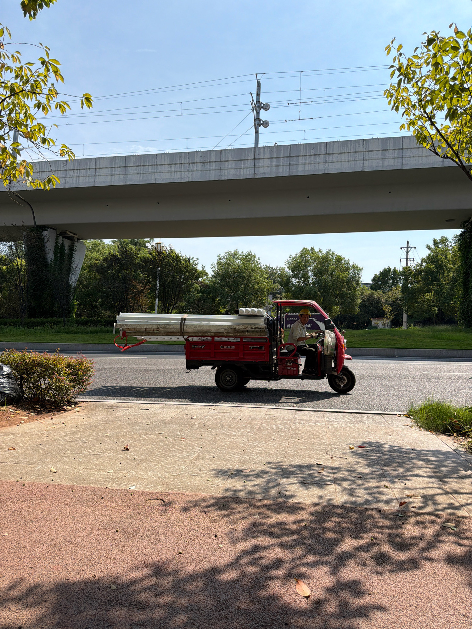 A red motorized tricycle with a canopy is driving on a road in Jiangjin, China. The tricycle is carrying a load of long, cylindrical objects, possibly pipes, secured with red straps. A man wearing a light-colored shirt and a yellow hard hat is operating the vehicle. The tricycle is passing under a large, elevated concrete structure, which appears to be an overpass or a railway bridge, with train tracks visible on top. The surrounding environment includes green trees and vegetation, a grassy median strip, and a sidewalk made of concrete and reddish paving stones. The sky is clear and blue. Shadows of trees are cast on the sidewalk.