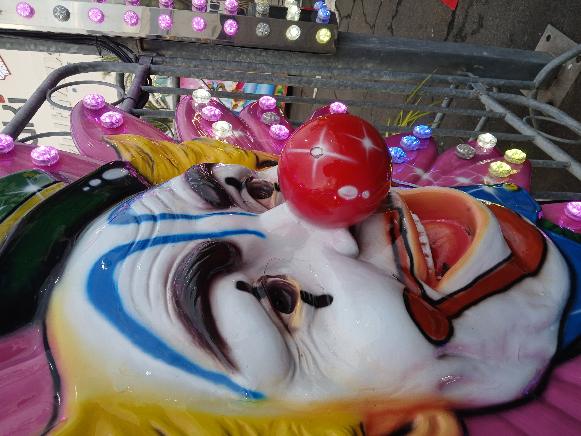The image displays a close-up, angled view of a large, static clown head, likely an element of an amusement park ride, positioned outdoors in Ciudad Guayana, Venezuela. The clown head features a white face, yellow textured hair, and bold makeup including blue vertical stripes on the forehead and cheeks, dark painted eyebrows, and black tear-like marks beneath the eyes. A prominent, glossy red spherical nose, approximately 6-8 inches in diameter, is centered on the face, exhibiting a distinct white starburst pattern. The mouth is open, revealing white teeth and a red interior, outlined with red and black paint. The lower portion of the head transitions into a blue and orange collar. To the right of the clown head, a weathered gray metal fence or barrier structure is visible, fitted with multiple circular LED lights that emit pink, white, yellow, and blue illumination. The ground beneath appears to be a dark paved surface. In the distant upper right background, a white building with partial black lettering and green foliage, consistent with palm trees, is discernible. No people are present in the frame.