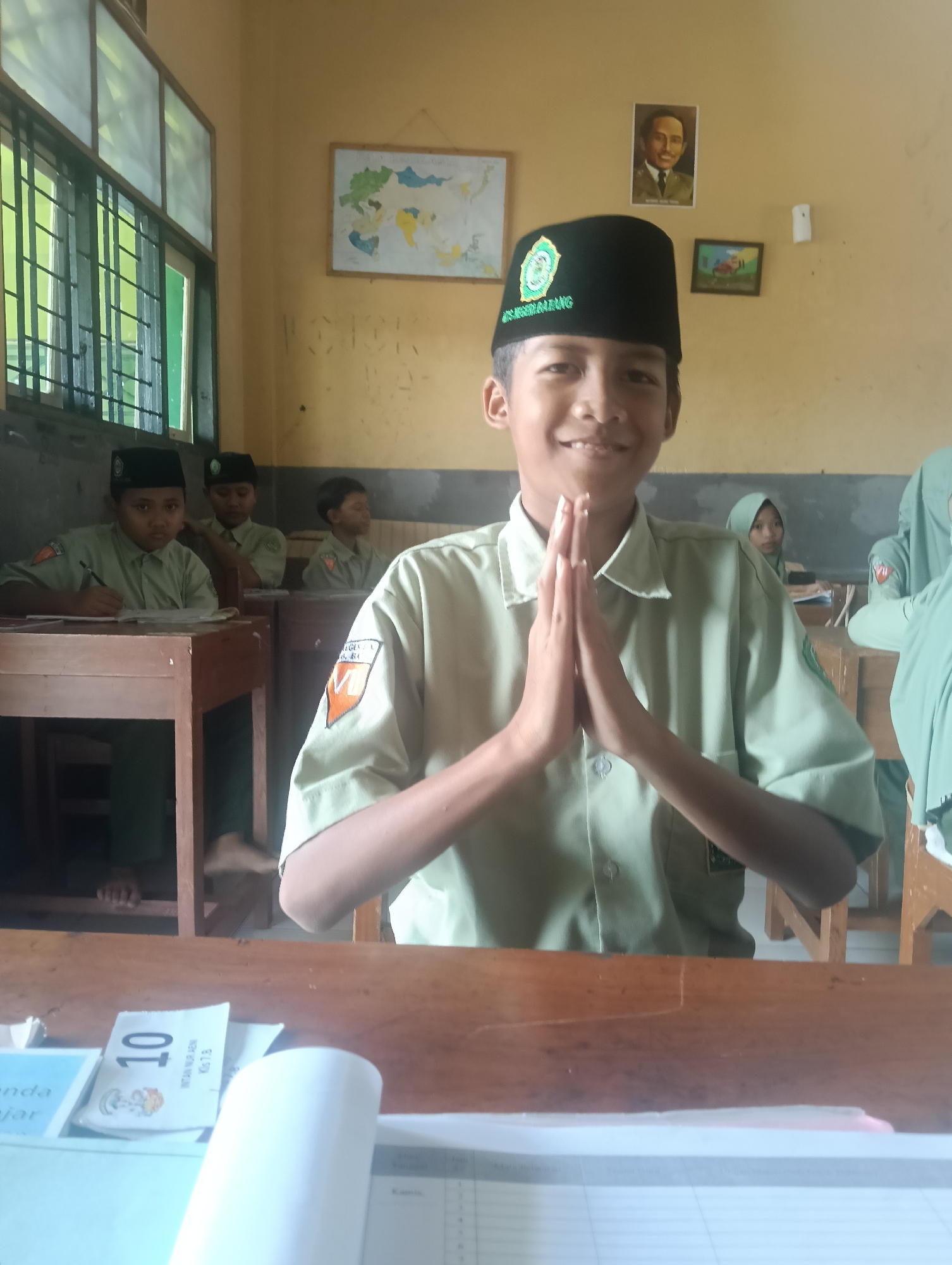 In Adinuso, Indonesia, a young boy in a black cap and a light green uniform smiles at the camera with his hands pressed together in front of his chest. He is in a classroom, seated at a wooden desk. Behind him, other students in similar uniforms are also seated at desks, some writing or looking forward. On the wall behind the boy, there are several framed pictures, including a portrait of a man in formal attire and a map. The classroom environment appears to be simple, with wooden desks, a large window with green bars, and a light-colored wall. The overall impression is of a school day in progress.