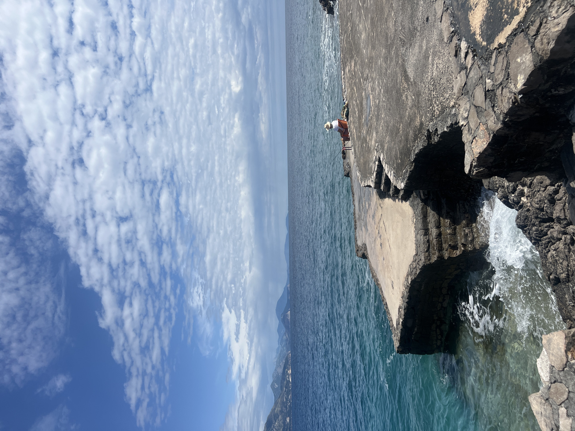 A pier made of concrete and rock extends into the Adriatic Sea on a sunny day in Prijevor, Montenegro. A lone man, wearing a white shirt, orange shorts, and a light-colored hat, fishes from the pier. The sea stretches into the distance under a sky filled with scattered white clouds. Mountains are visible on the distant horizon. Waves crash against the rock base of the pier.