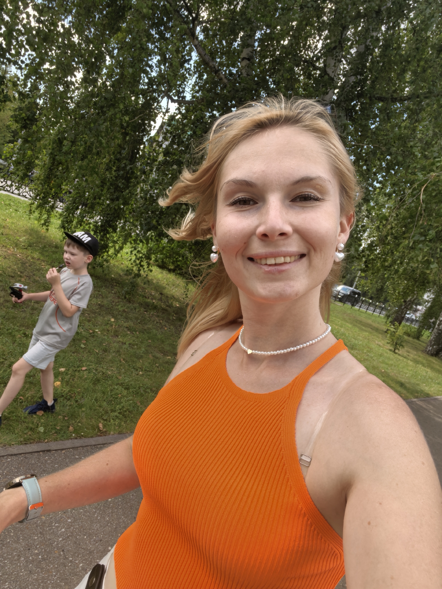 In this selfie, a blonde woman with fair skin and brown eyes smiles at the camera. She is wearing a bright orange ribbed tank top, a pearl necklace with a small gold heart pendant, and white heart-shaped pearl earrings. A tan line is visible on her left shoulder. Her hair is windswept. In the background, a young boy wearing a grey t-shirt, light blue shorts, and a black baseball cap with "AM" on it, is standing on a grassy slope. He is holding a small black object in his right hand and appears to be interacting with something in front of him. Behind the boy, a path and more trees are visible. The overall scene suggests an outdoor setting in a park or recreational area. The image was taken on a bright day, likely in a temperate climate given the foliage. Without further context, it is impossible to definitively determine the specific location in Sterlitamak, Russia.
