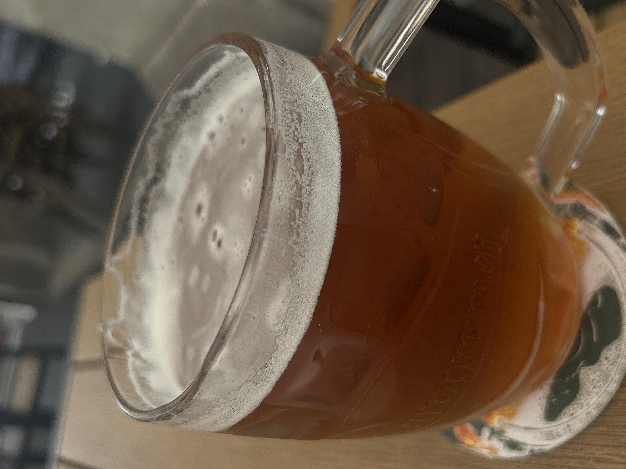 A close-up, high-angle shot captures a glass beer mug filled with a reddish-brown beverage, likely a lager or ale, resting on a light-colored wooden table. A thick layer of white foam tops the drink, and "lacing" from the foam adheres to the inner wall of the clear glass mug. The mug features a prominent handle on the right side. Beneath the mug is a circular coaster with an indistinct, partially obscured pattern, including a dark, irregular shape. Faintly embossed or printed on the side of the glass, the word "Prazdroj" is visible, rotated, suggesting a connection to Pilsner Urquell, a prominent Czech beer brand. The background is softly blurred, showing only vague shapes, possibly shelves or other indoor elements. The image is taken indoors, with no specific time of day or weather discernible. The overall scene suggests a casual setting, possibly a pub, restaurant, or home, consistent with the given context of Liberec, Czechia.