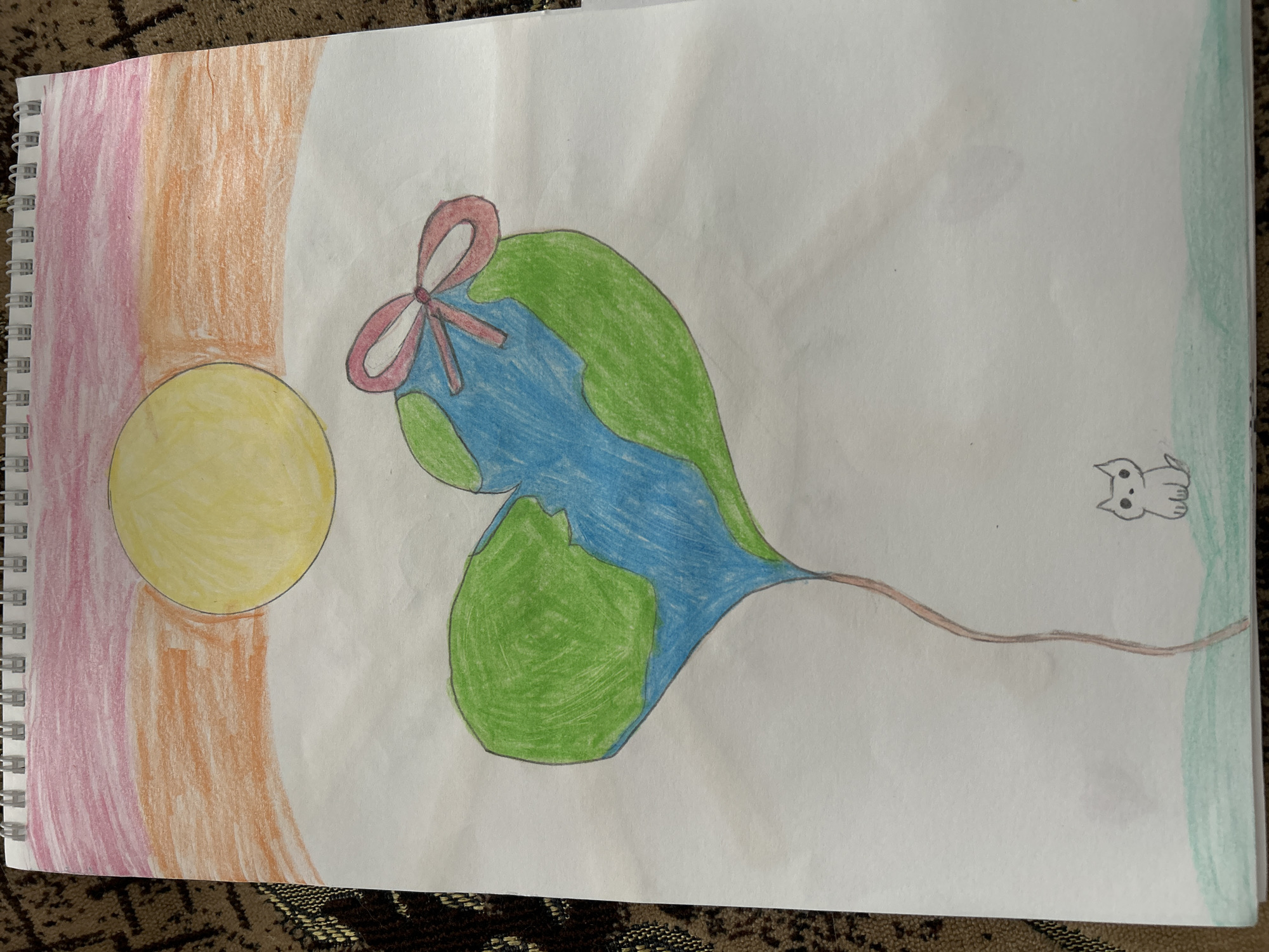 The image is a child's drawing on a page from a spiral-bound notebook. On the left side of the drawing, there is a yellow circle with orange and pink shading behind it. To the right of the circle is a stylized heart-shaped object colored with blue and green, with a red bow at the top. A thin brown line extends from the bottom point of the heart-shaped object to the right edge of the paper. Near the right edge of the paper is a small, simplified drawing of a cat. The background is mostly white, with a light green border along the right edge. There is no indication of Uray, Russia, or any other specific location.