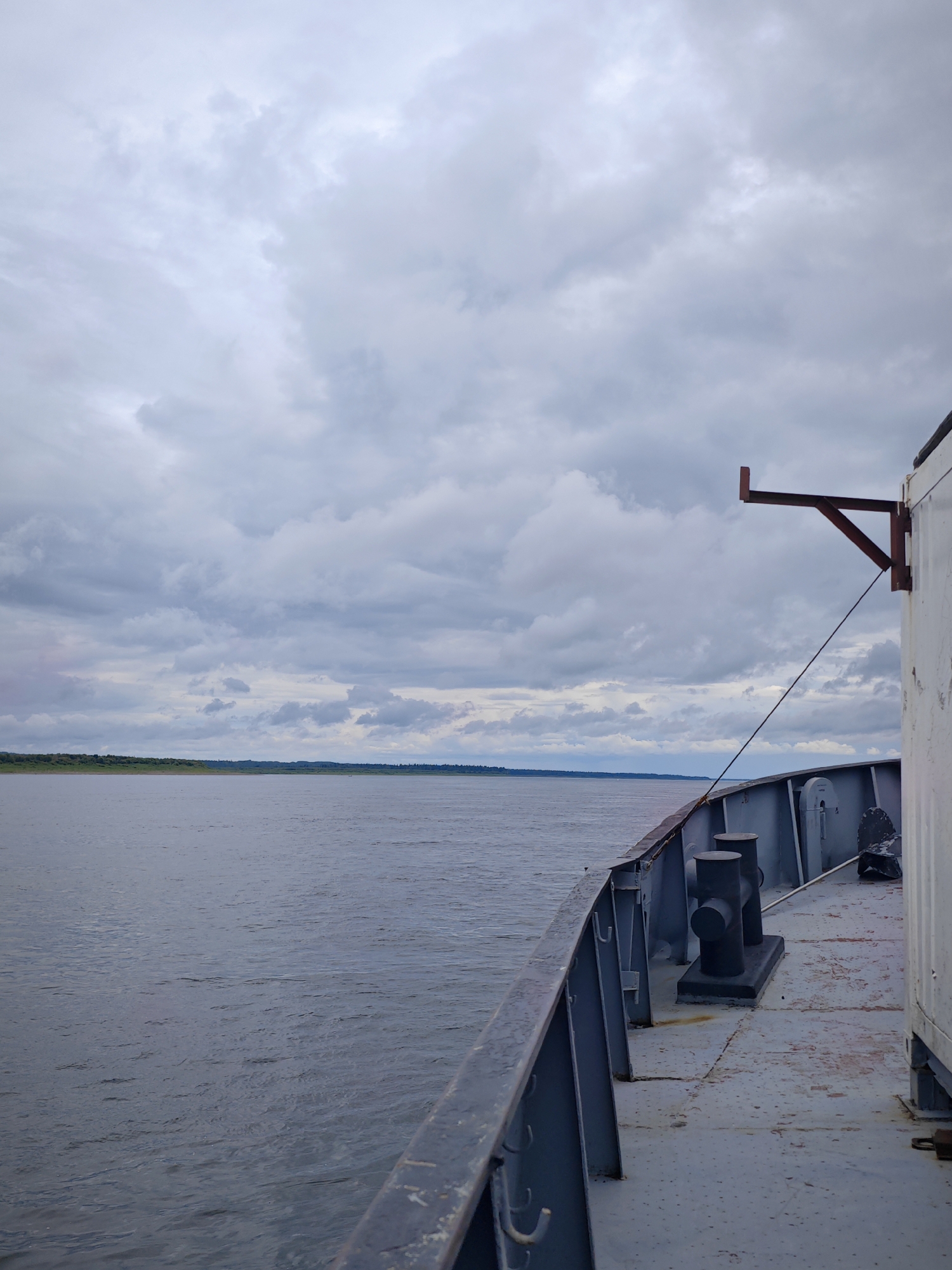 The image shows a view from the deck of a ship on a river in Zotino, Russia. The sky is overcast with gray clouds. The river is wide and calm, with a tree-lined shore in the distance. On the deck of the ship, there are bollards and railings. A white cargo container is visible on the right side of the image. The scene is likely captured during daytime, given the ambient light. There are no people or animals visible in the image.