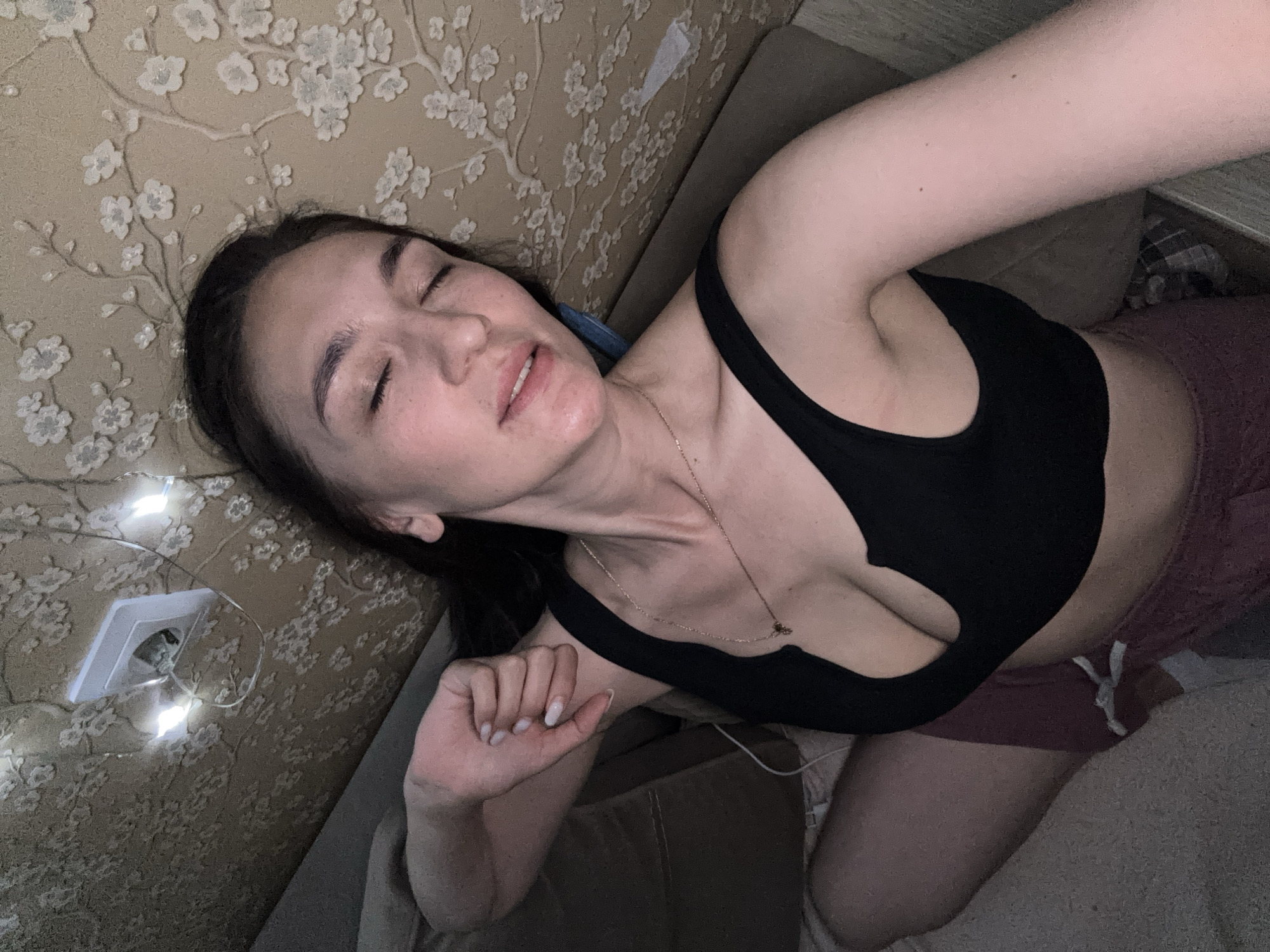 A woman with dark hair lies on a couch in a room with patterned wallpaper. She is wearing a black sports bra and maroon shorts. A string of lights is visible near an electrical outlet on the wall. The location is not specified, and there is no information available about it being in Sochi, Russia.