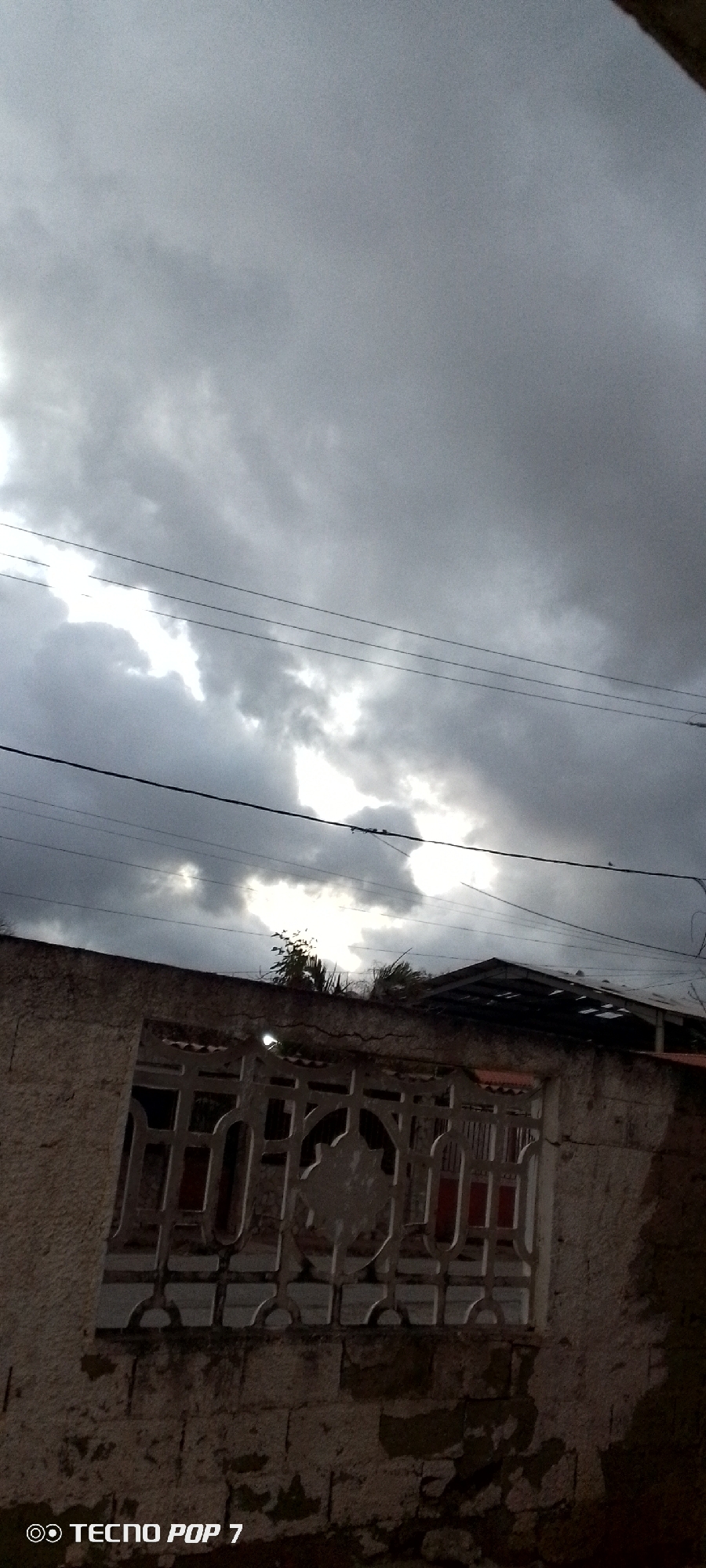 The image captures an overcast sky in what appears to be Altagracia, Venezuela. Heavy, dark clouds dominate the upper two-thirds of the frame, partially obscuring the sun, which peeks through as a hazy, bright patch. Several electrical or telephone wires stretch horizontally across the middle of the image. In the bottom third, there's a weathered concrete or stone wall topped with a decorative metal grate. Behind the wall, the roof of a building is visible, with parts of the roof appearing damaged or deteriorated. A small tree or plant grows just behind the wall. The lower-left corner has a visible "TECNO POP 7" watermark. The overall lighting is subdued, casting the scene in shades of gray. The foreground wall shows signs of aging, with cracks and discoloration evident.