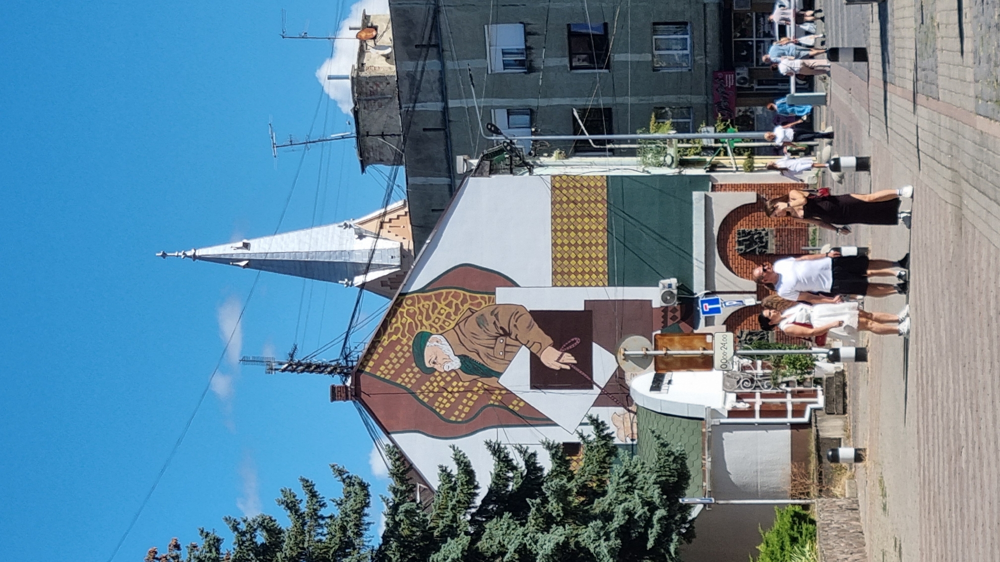 This image depicts a street scene in Mukachevo, Ukraine, featuring a large mural on the side of a building. The mural is a portrait of an elderly man wearing a green beret and a brown jacket, holding a string of beads. He is depicted against a backdrop of abstract patterns. In the background, a tall, silver church spire is visible above the rooftops.
In the foreground, several people are standing on a sidewalk or pedestrian crossing. Three individuals are most prominent: a woman in a white dress, a man in a white t-shirt and black shorts, and a woman in a long black dress. They appear to be conversing or waiting. Behind them, other pedestrians are walking along the street, and commercial buildings line the opposite side. The environment suggests a sunny day with a clear blue sky, characteristic of an urban setting.