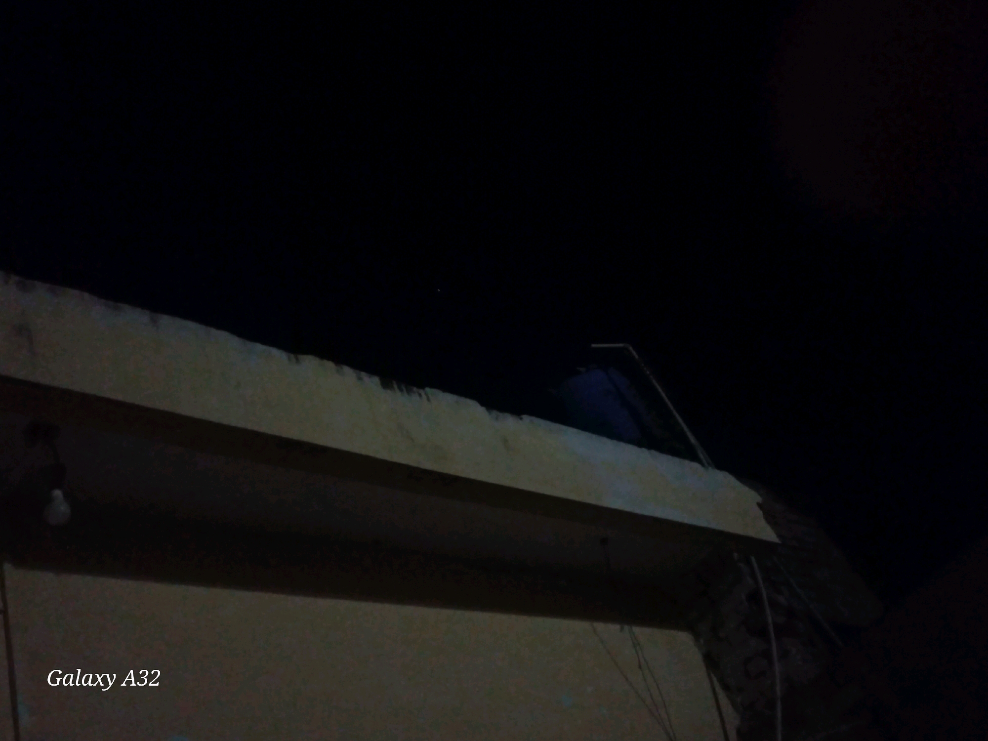 This is a nighttime photograph taken outdoors. The foreground features a portion of a building, likely a wall or rooftop, with a visible overhang. A light fixture with a bulb is attached to the underside of the overhang on the left side of the frame. The background consists of a dark sky with a single, faint star visible in the upper center. The words "Galaxy A32" are watermarked in the lower left corner of the image. The scene appears to be in Dalasa, Pakistan, based on the watermark, suggesting a residential or urban setting.