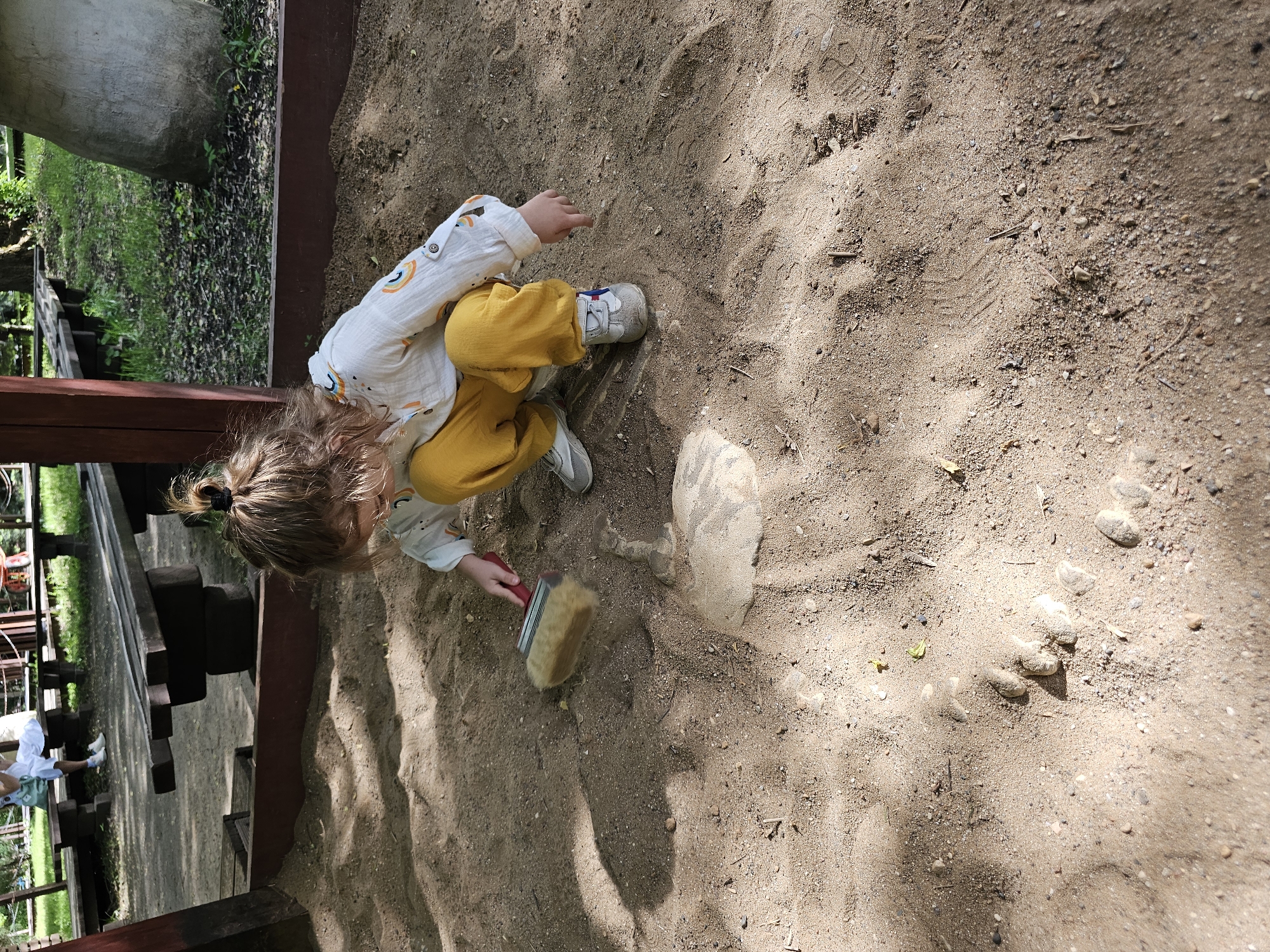 A child, with blonde hair pulled back in a ponytail, is playing in a sandbox. The child wears a long-sleeved, white shirt with rainbow designs and yellow pants. The child is kneeling and holding a tan-colored brush with a red handle. There is an object half-buried in the sand next to the child. The sandbox is bordered by dark wood. Shadows from trees fall across the sand. The location is potentially Khimki, Russia.